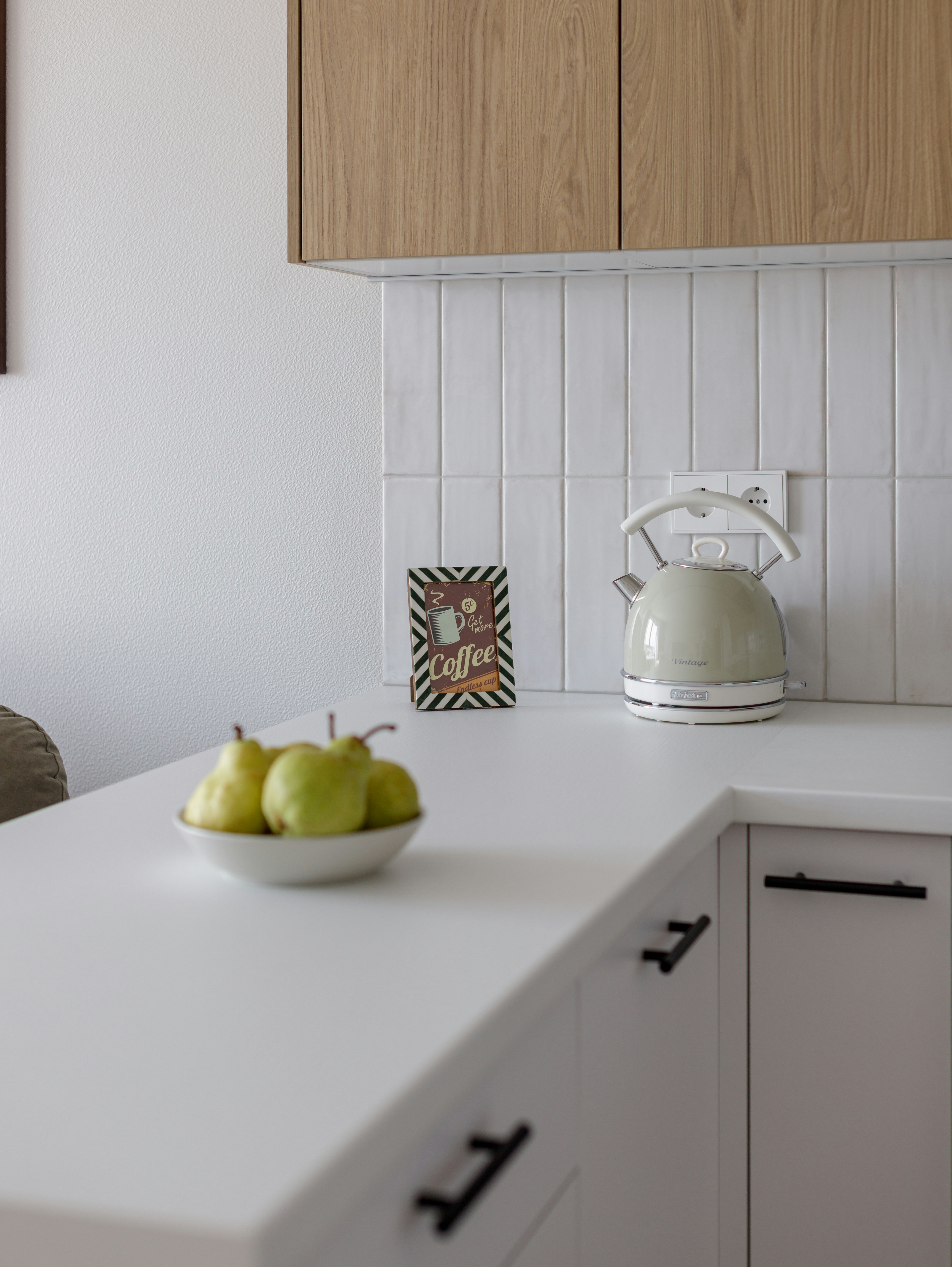 A bowl of green pears beside a kettle on a white kitchen counter