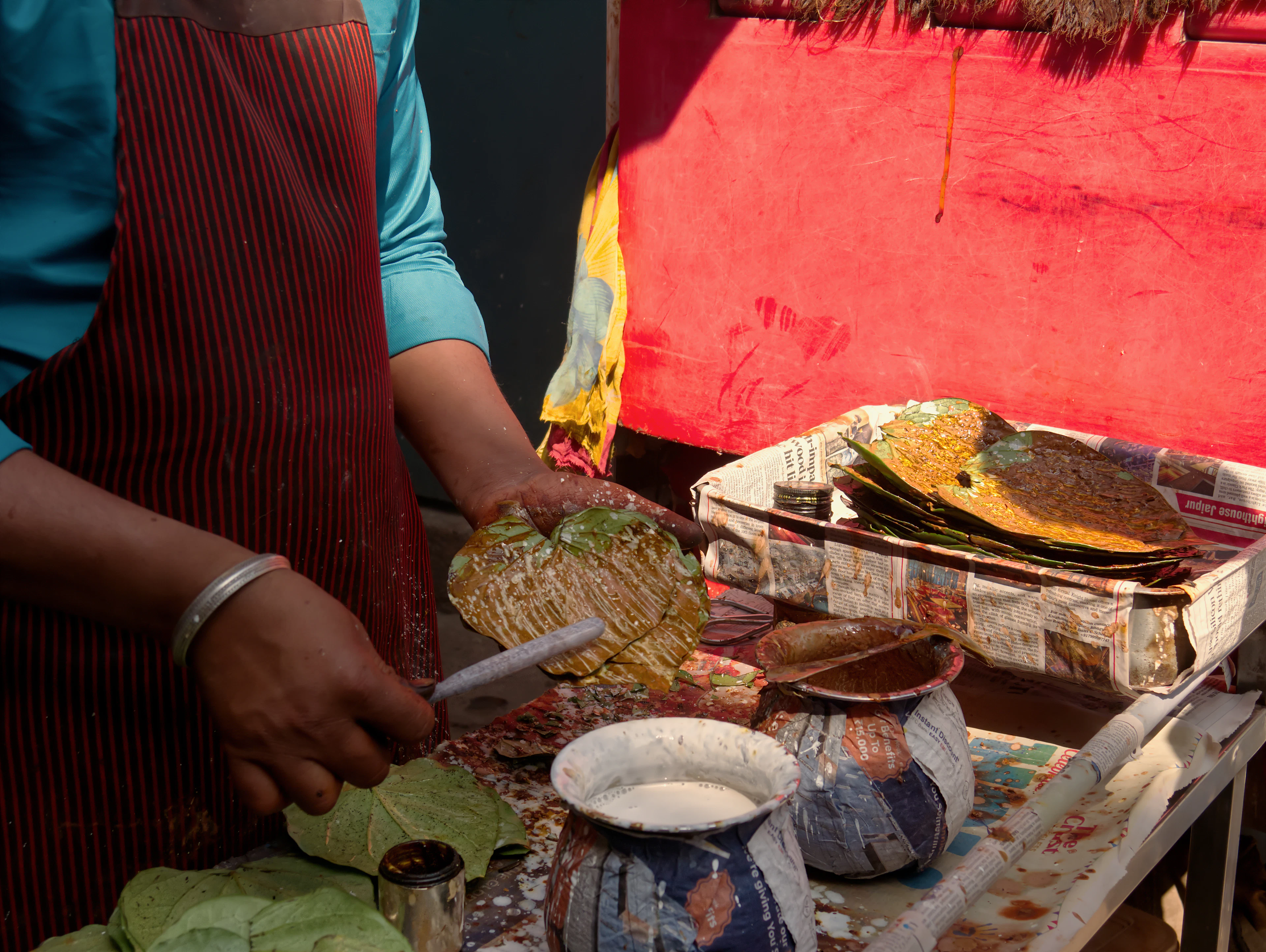A paan seller at a roadside stall spreading white lime paste across a fresh green betel leaf with a small blade, a tray of dressed leaves and pots of fillings on the counter beside her.