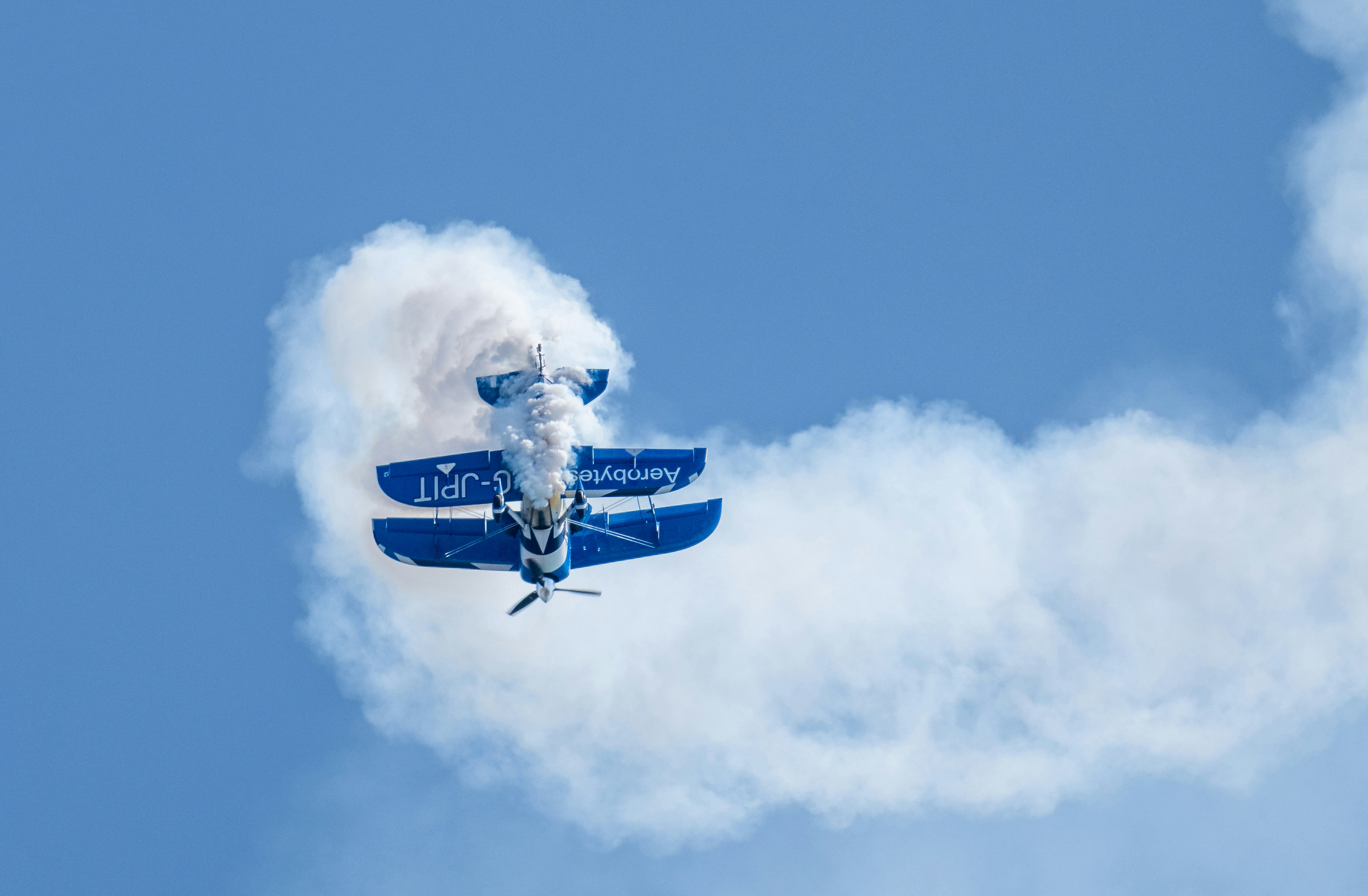 A blue biplane performing an aerial maneuver with a thick trail of white smoke