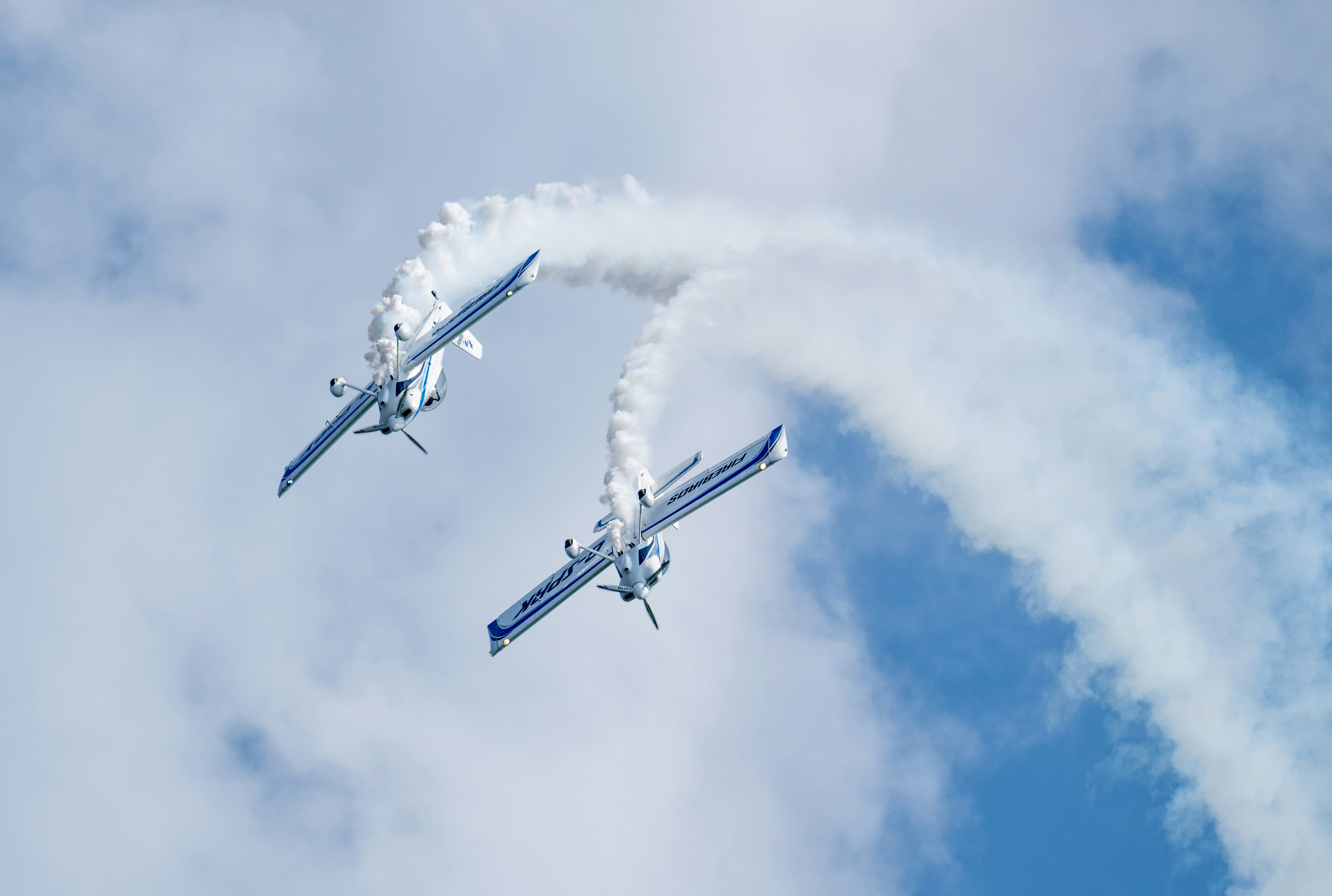 Two white stunt planes performing aerial maneuvers with smoke trails against a blue sky