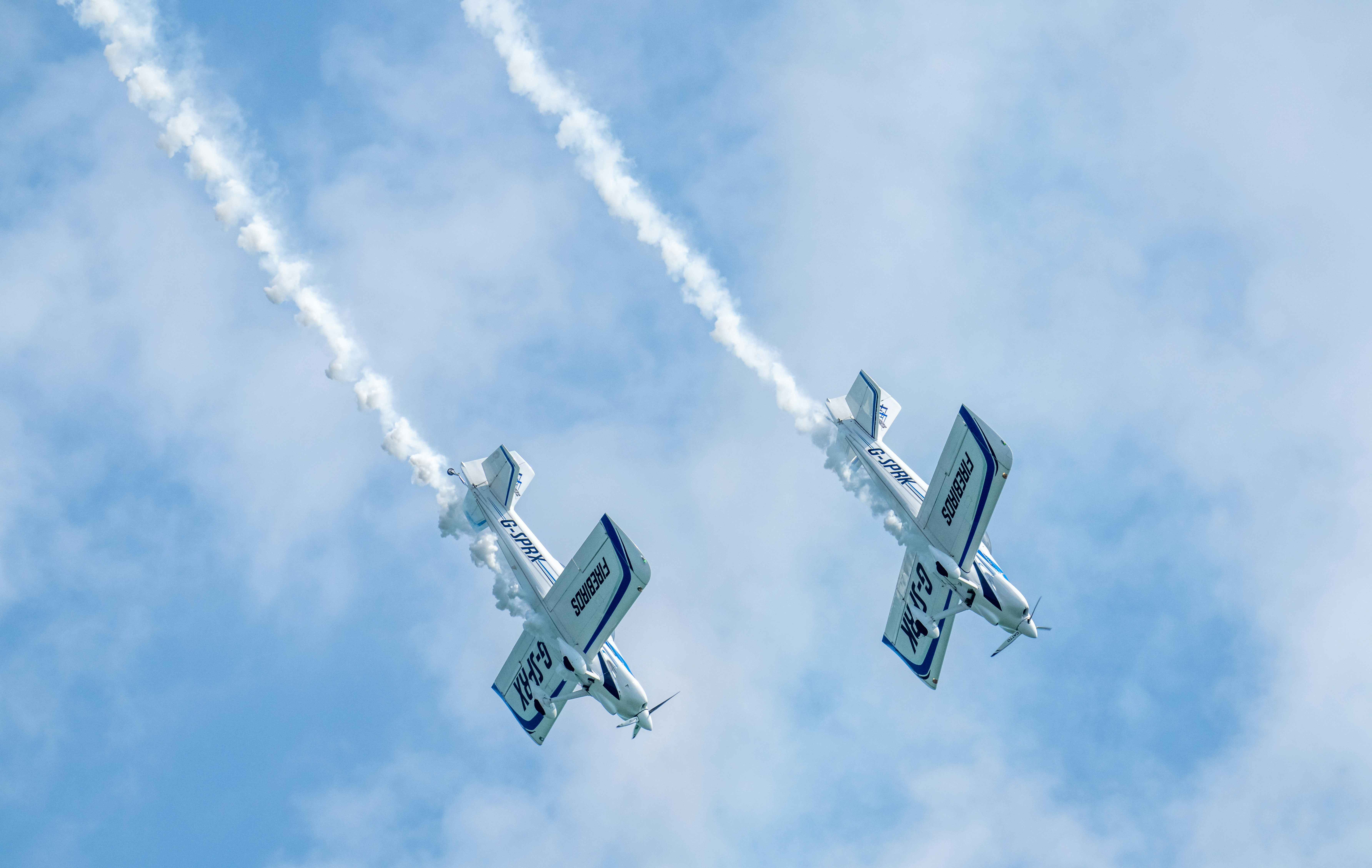 Two white aerobatic planes flying in formation against a blue sky with smoke trails