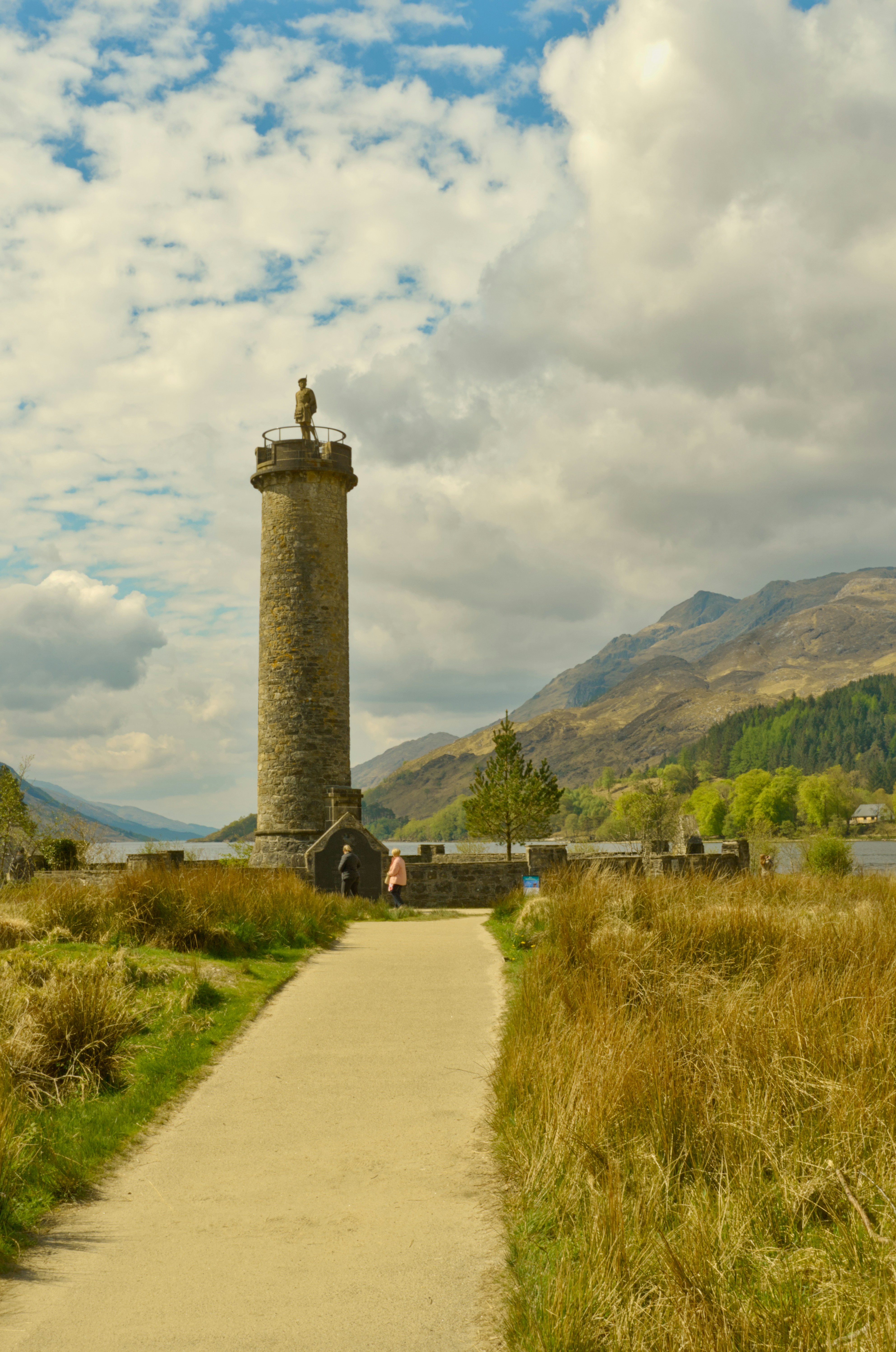 A stone monument tower with a statue on top near a lake and mountains