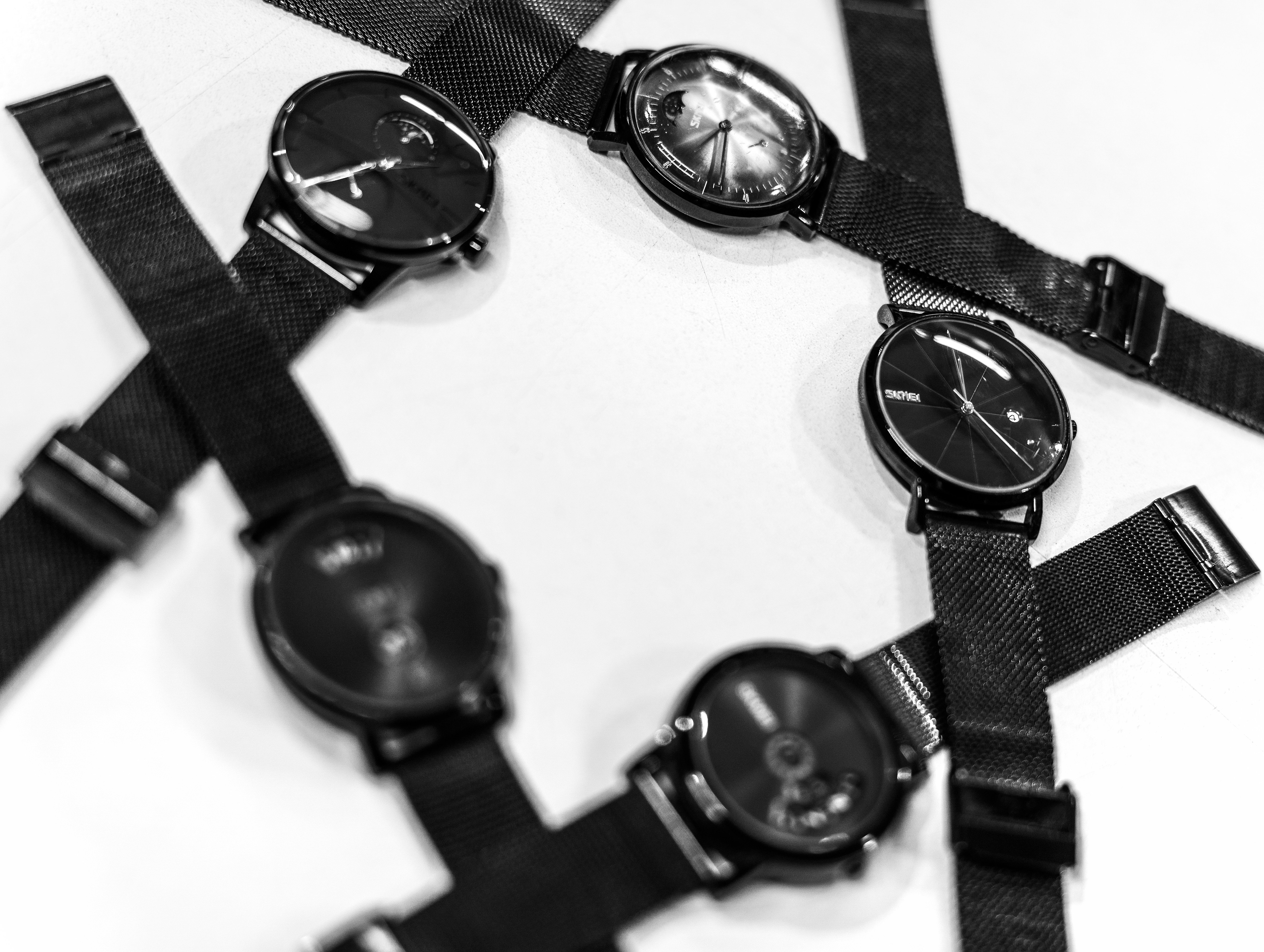 Four black wristwatches with mesh bands arranged in a circle on white