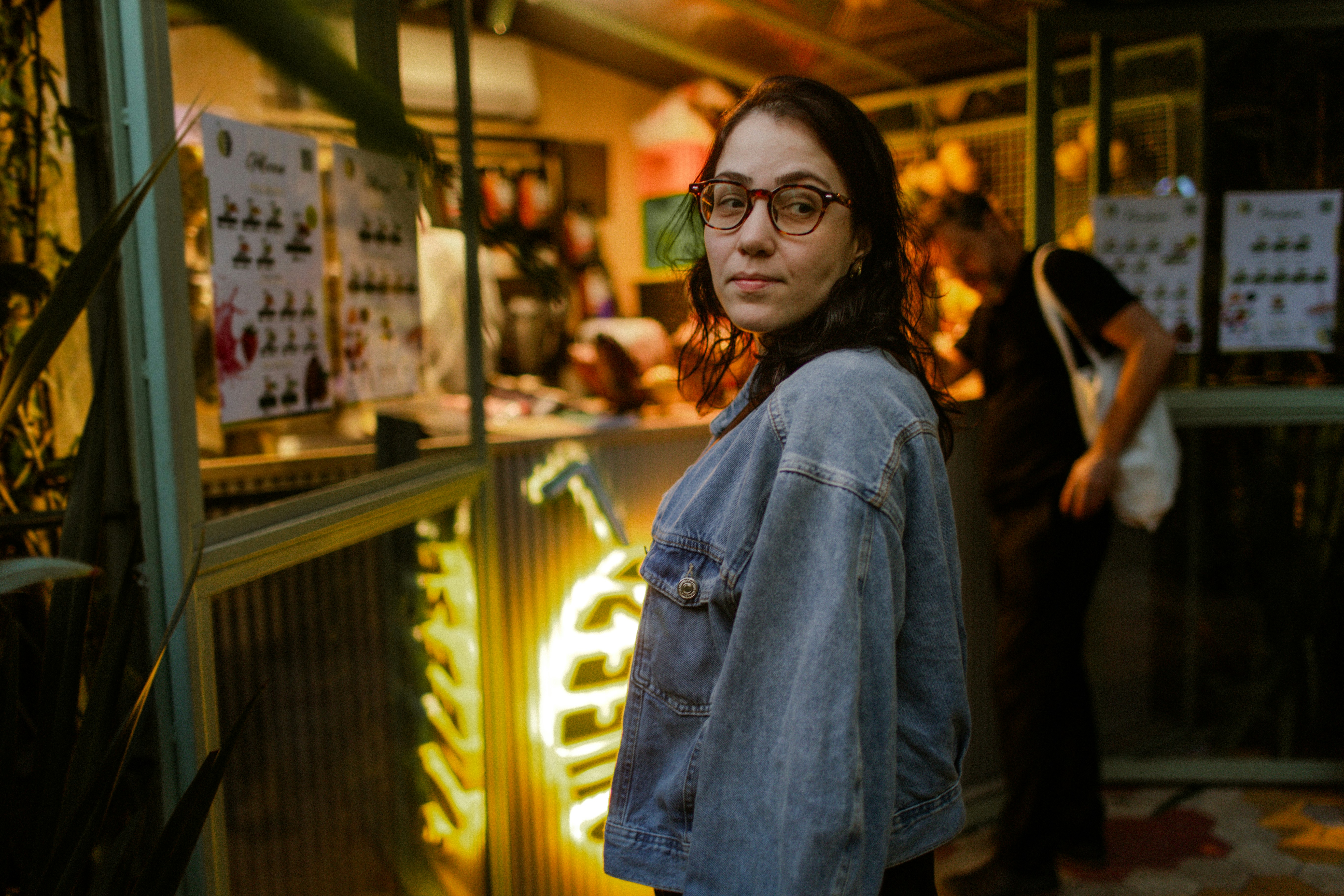 A woman in a denim jacket and glasses looking back in a neon-lit cafe