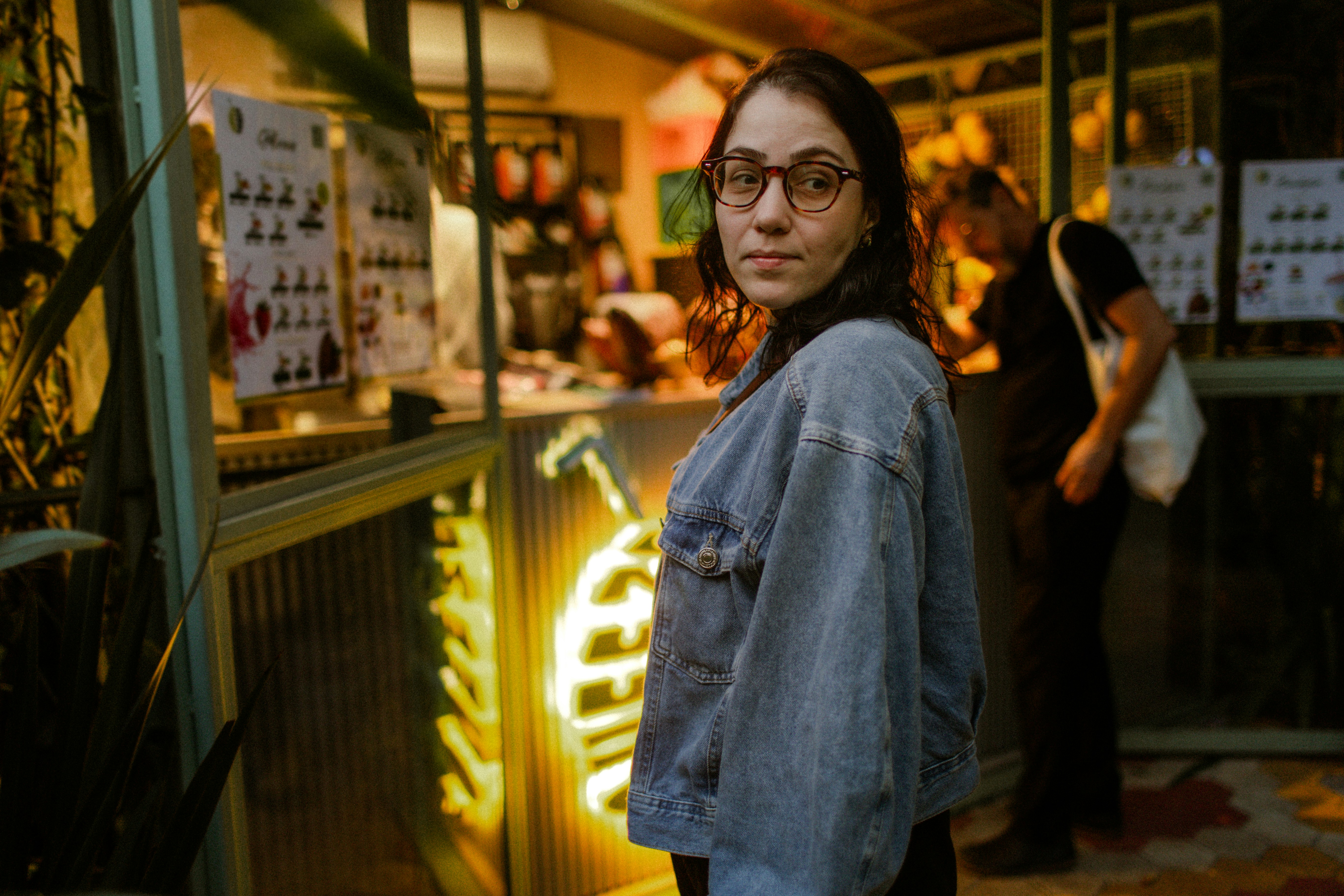 A woman in a denim jacket and glasses looking back in a cafe