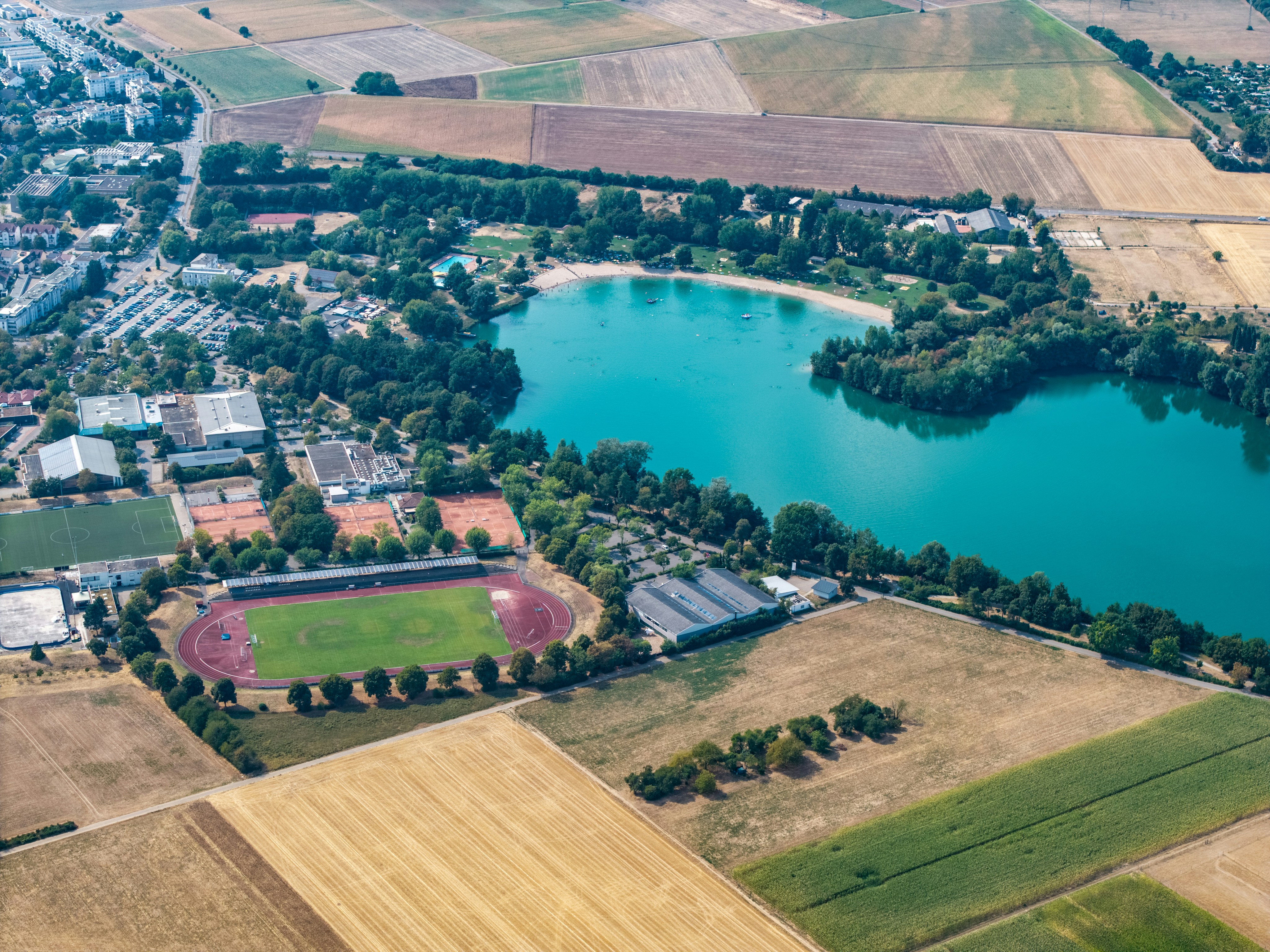 Aerial view of a turquoise lake surrounded by trees, sports fields, and farmland