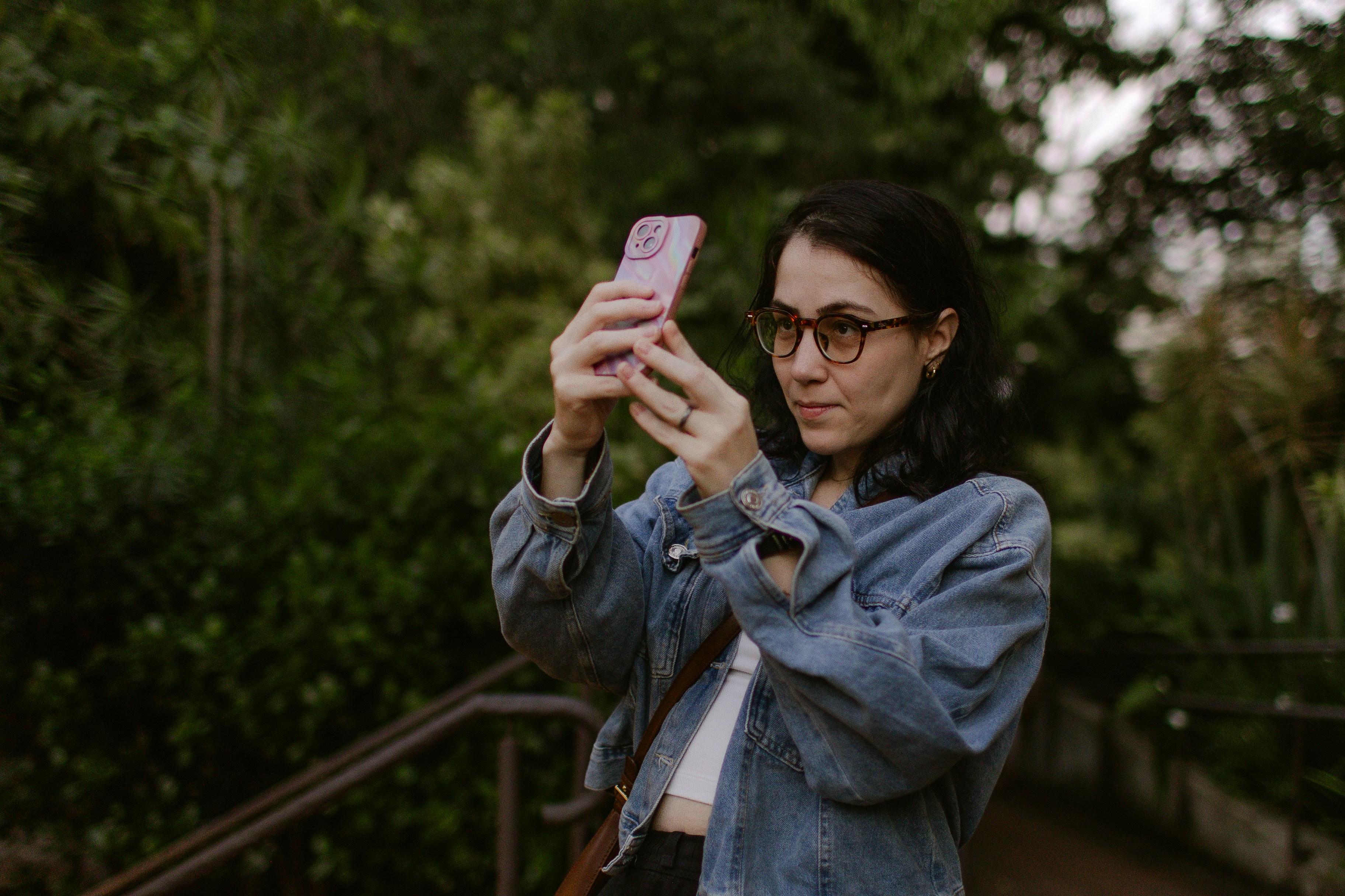 A woman in a denim jacket taking a photo with a pink smartphone outdoors