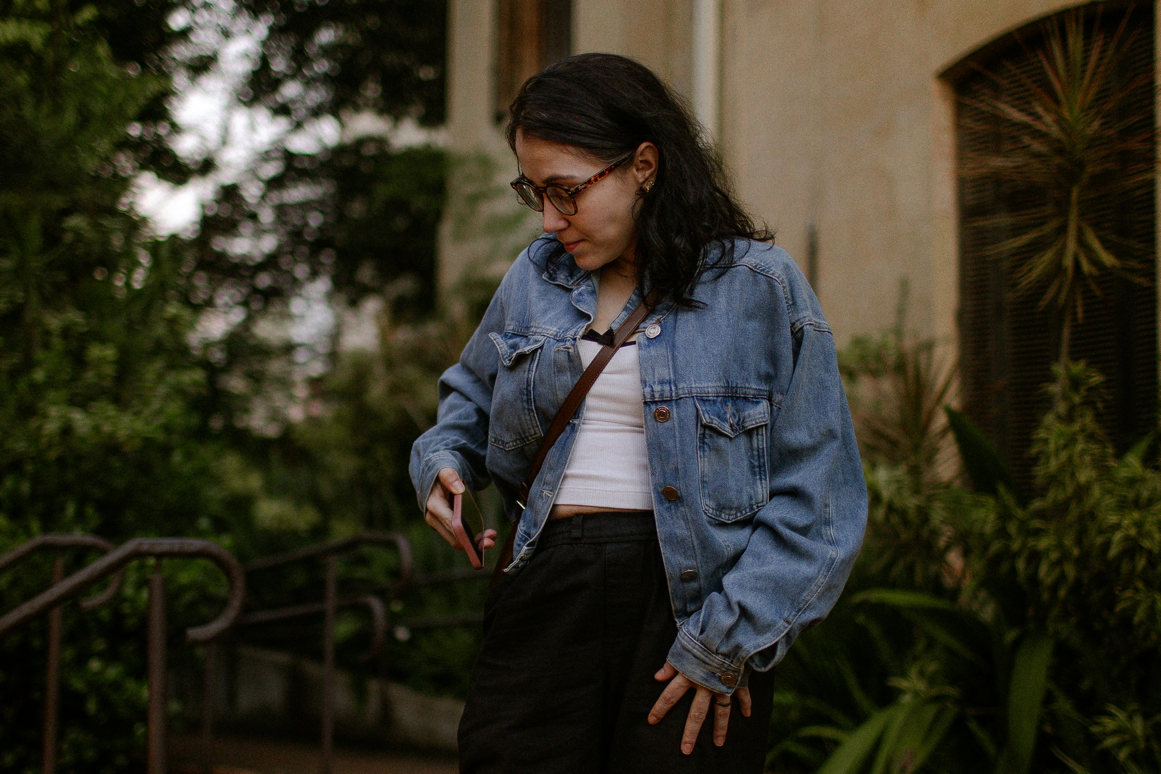 A woman in a denim jacket holding a smartphone outdoors near green foliage
