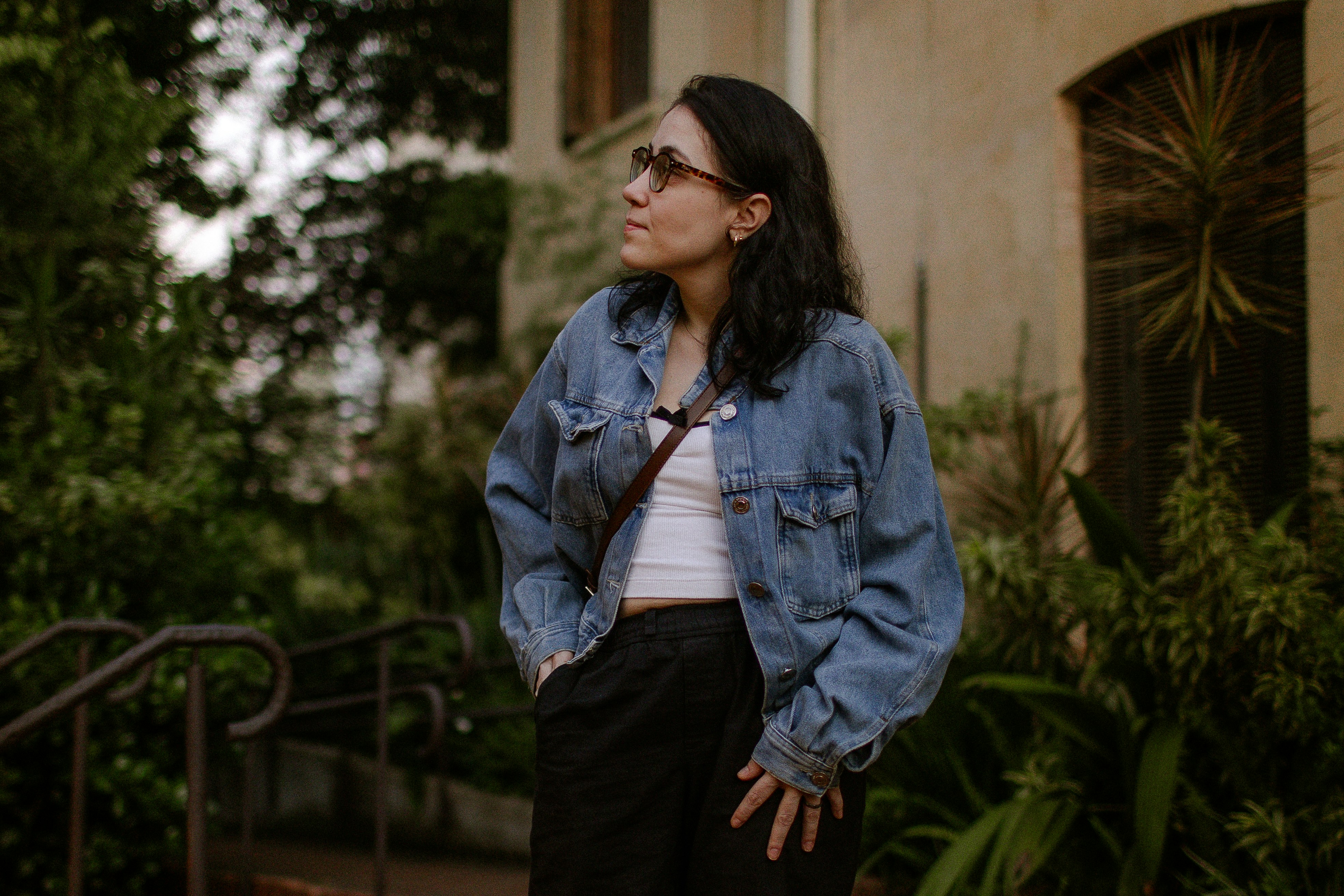 A woman in a denim jacket and white top standing outdoors near greenery