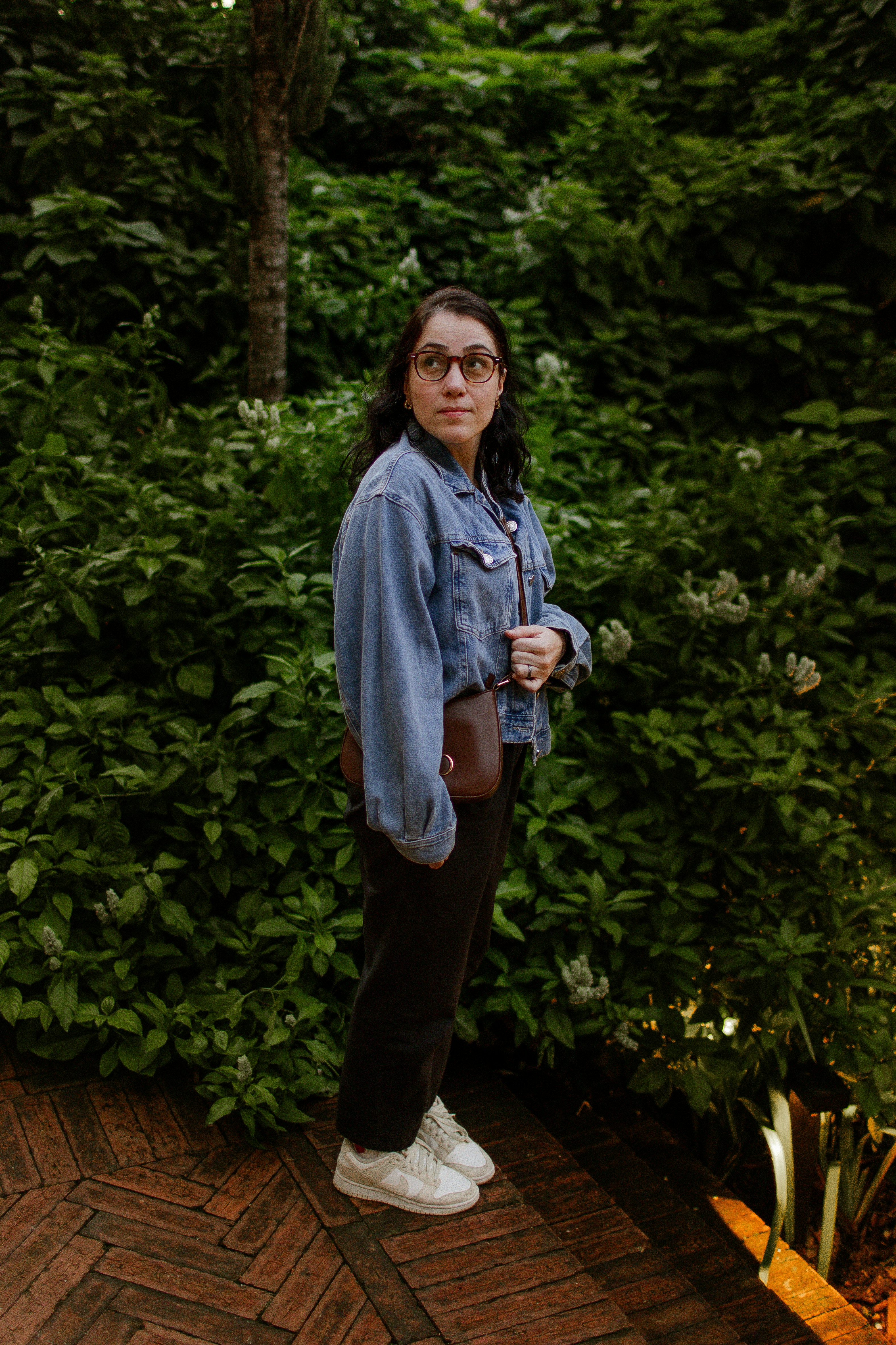A woman in a denim jacket and glasses standing before lush green foliage