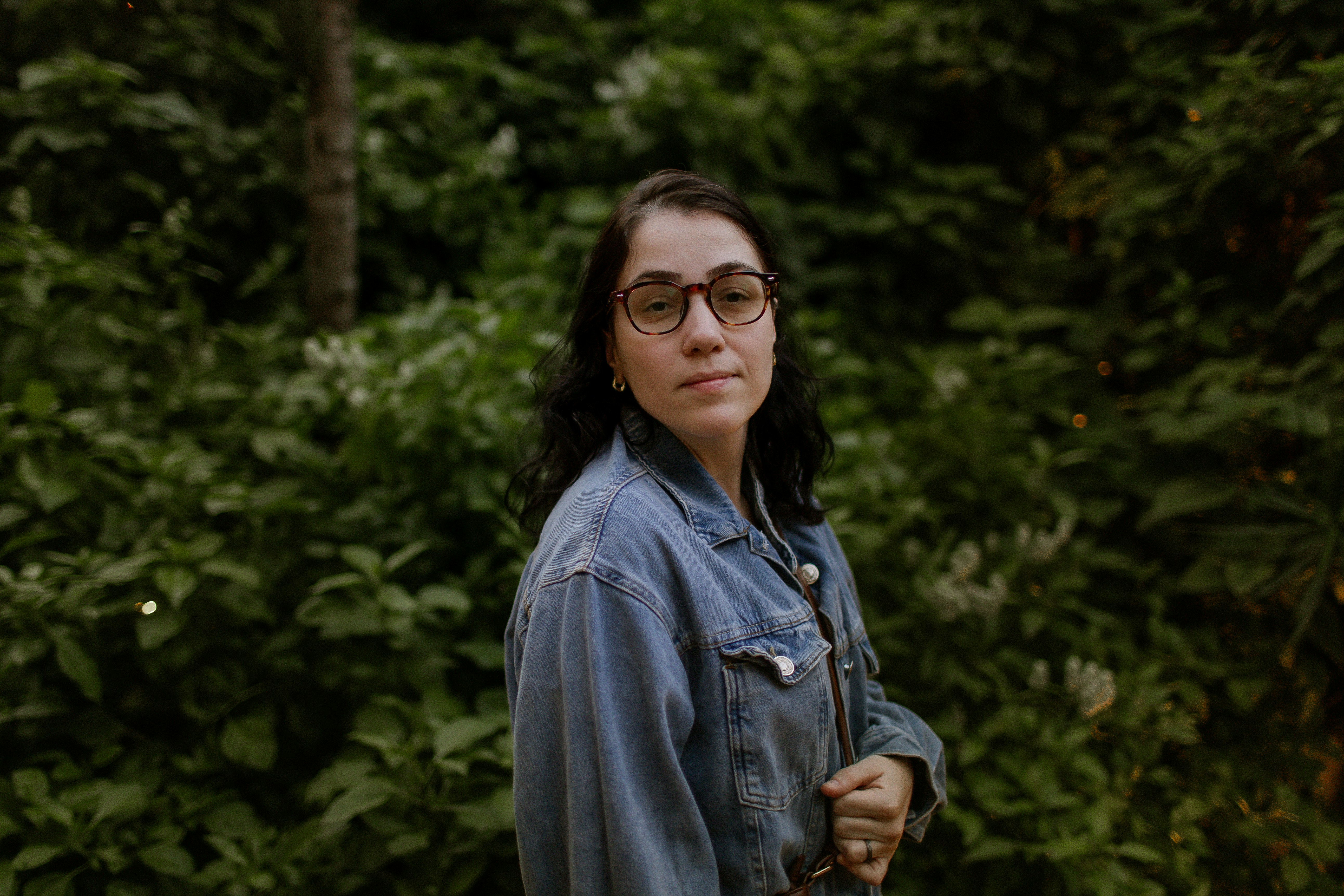 A woman with dark hair and glasses wearing a denim jacket in a garden