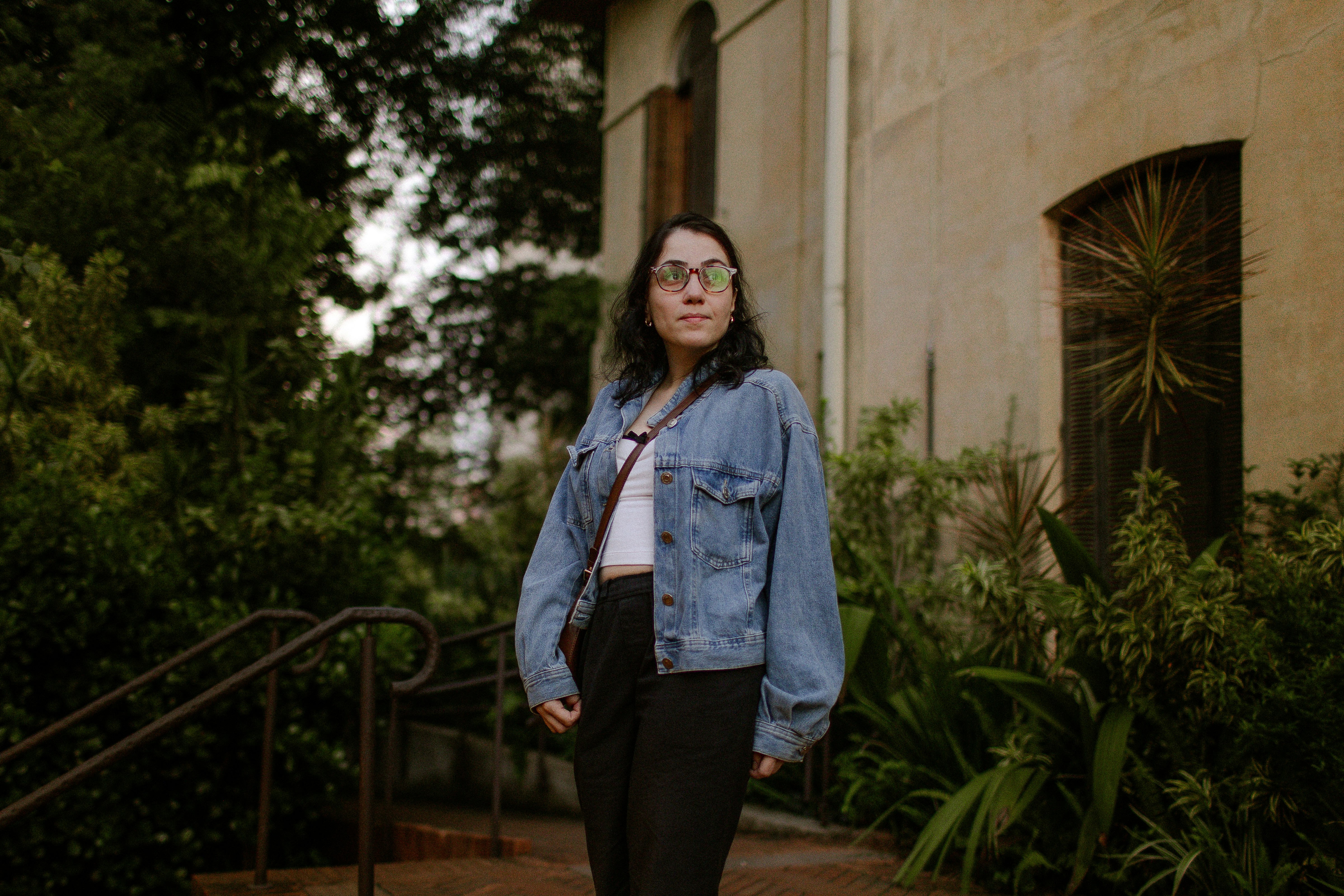 A woman in a denim jacket and glasses outdoors near a beige building