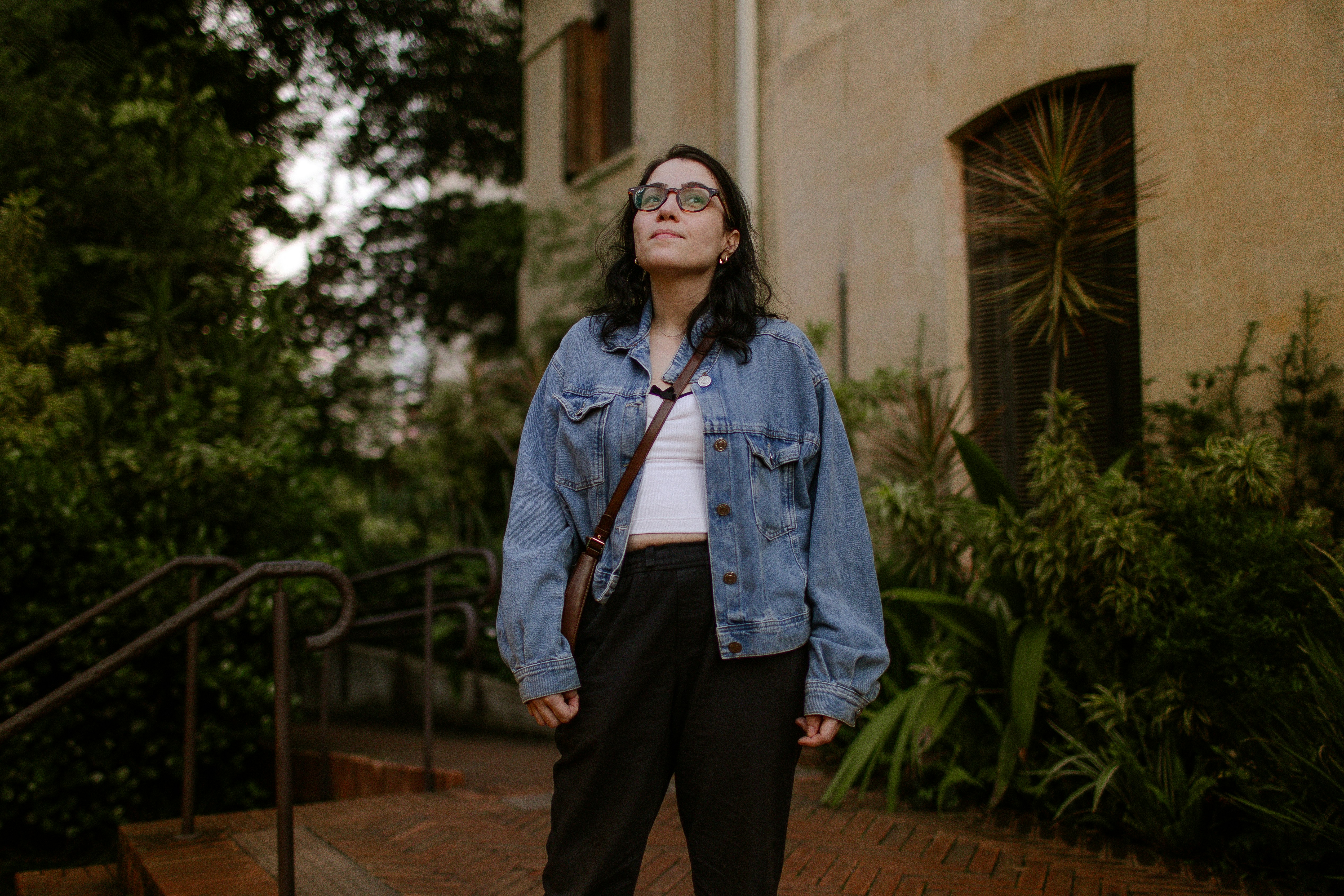 A woman in a denim jacket and glasses looking up in a garden