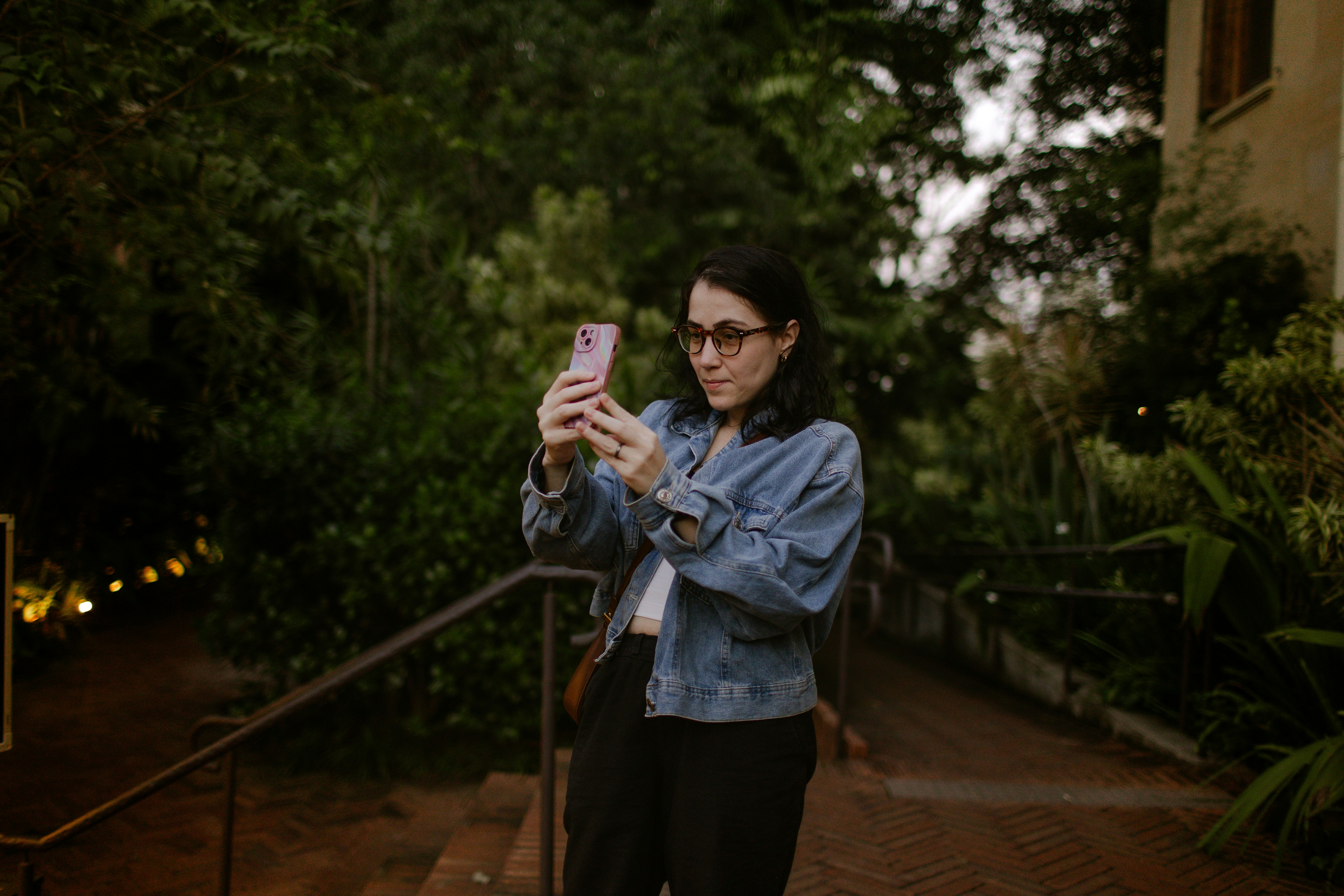 A woman in a denim jacket taking a photo with her smartphone outdoors