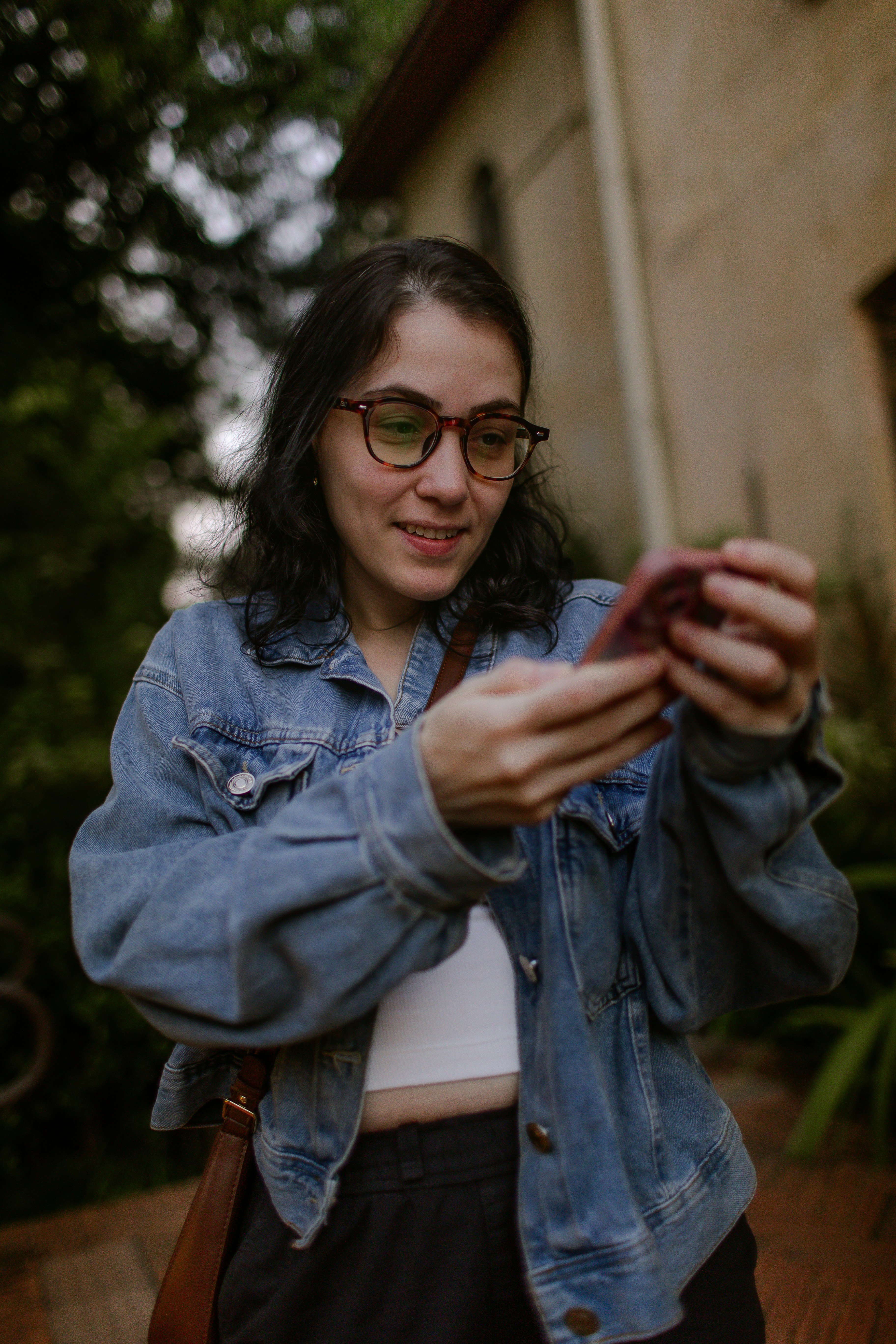 A woman with glasses wearing a denim jacket looking at her smartphone outdoors