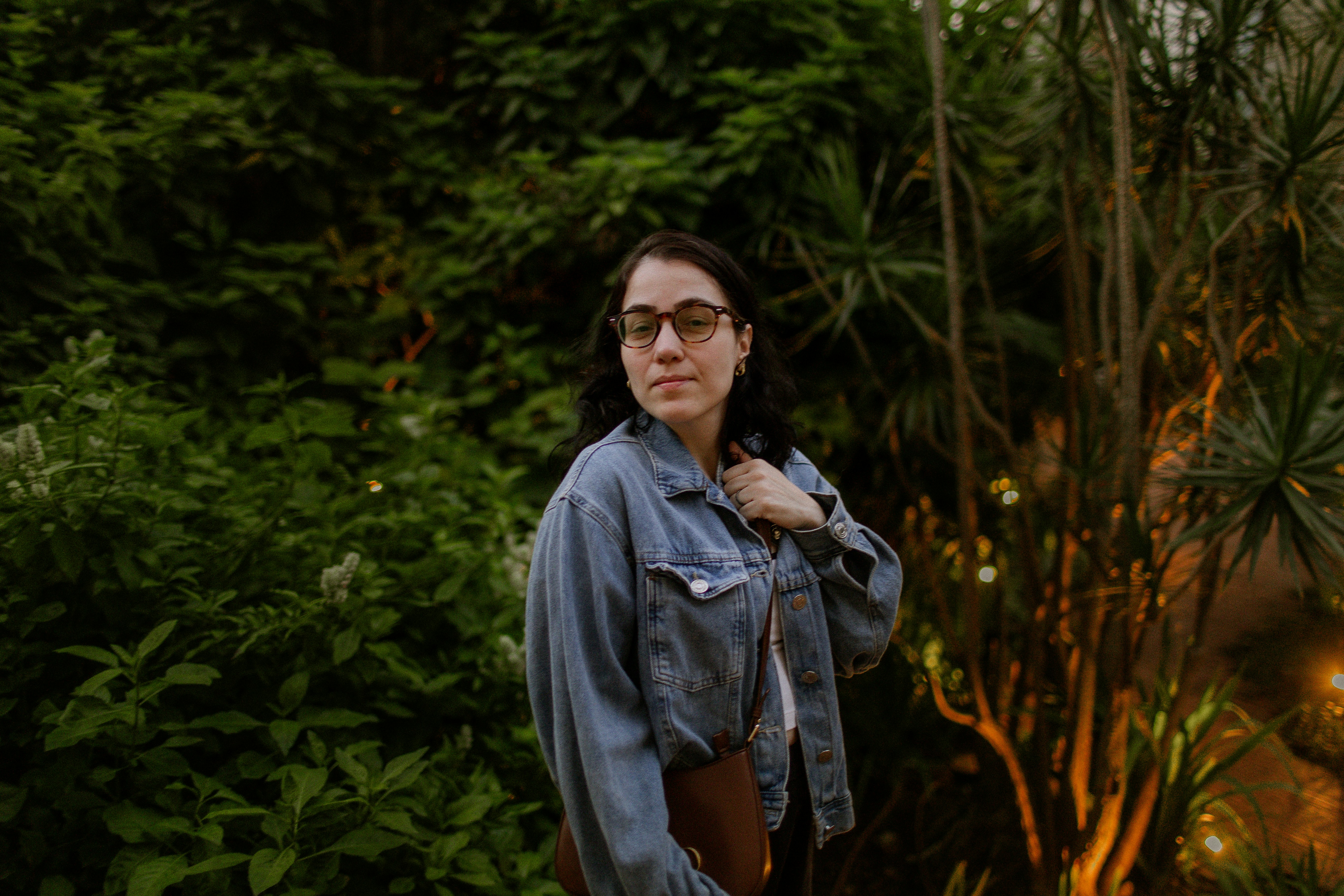 A woman with glasses and a denim jacket in a lush garden