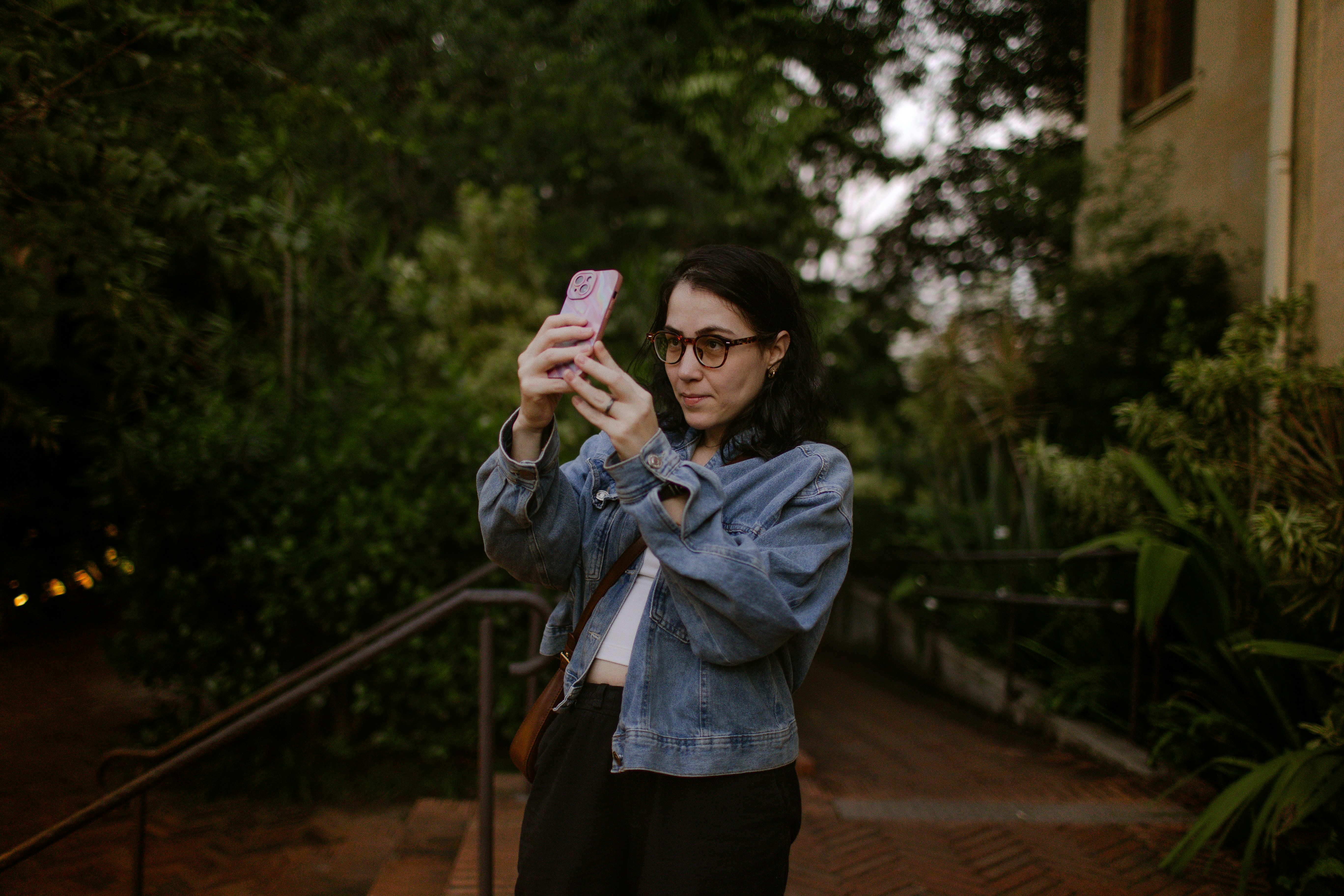 A woman in a denim jacket taking a photo with a pink smartphone outdoors