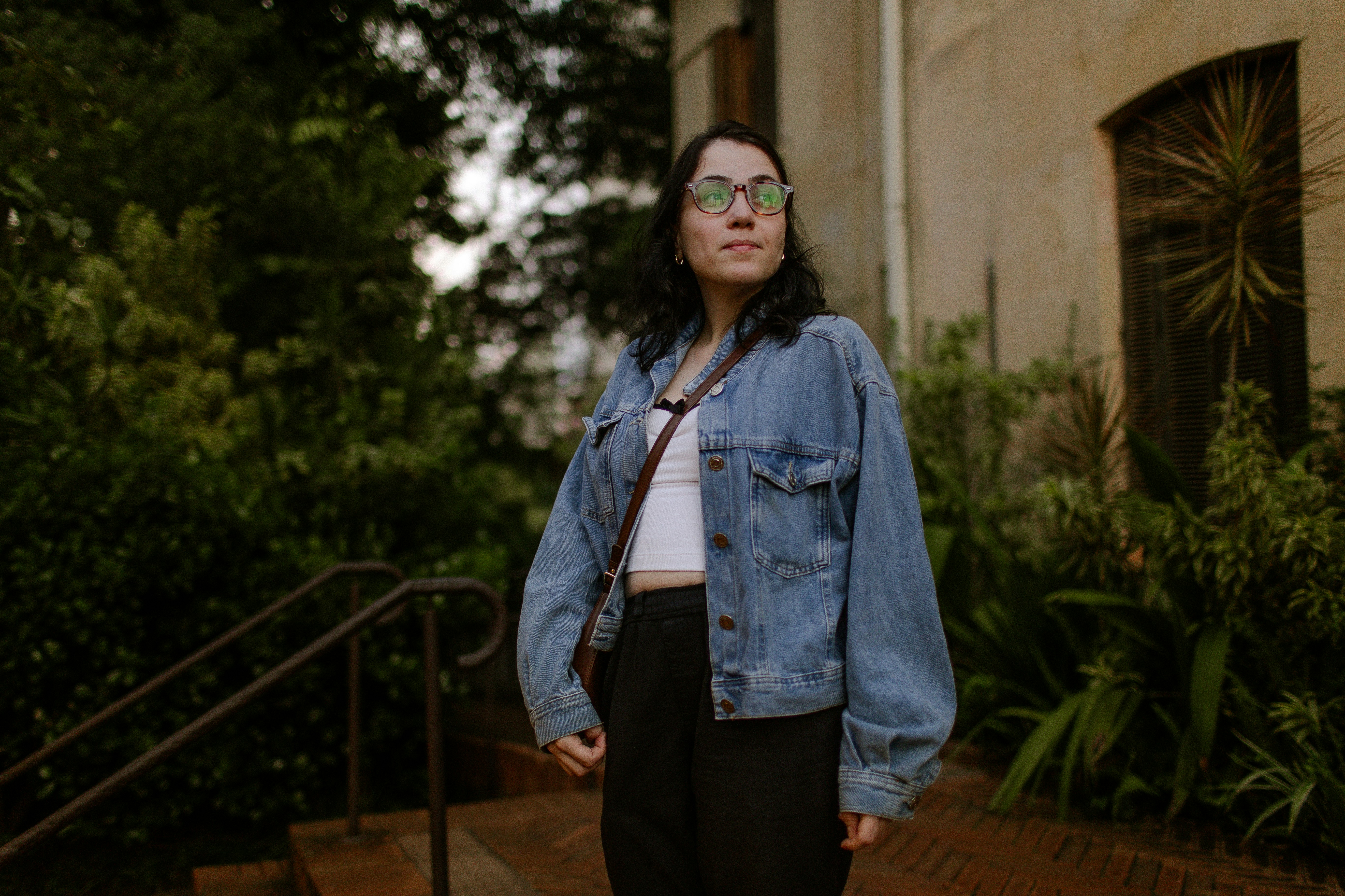 A woman in a denim jacket and glasses standing outdoors near lush greenery