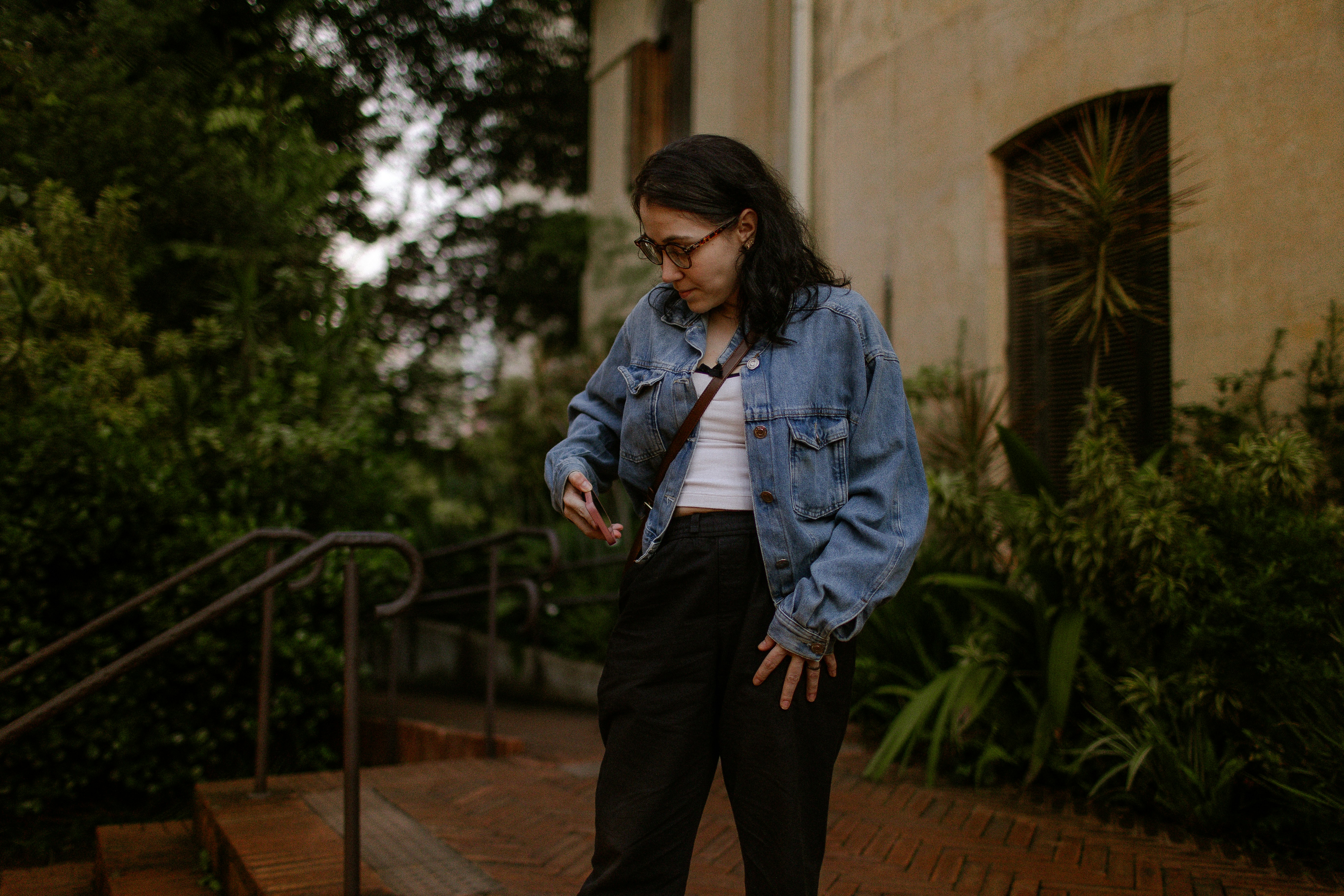 A woman in a denim jacket and glasses looking at her phone outdoors