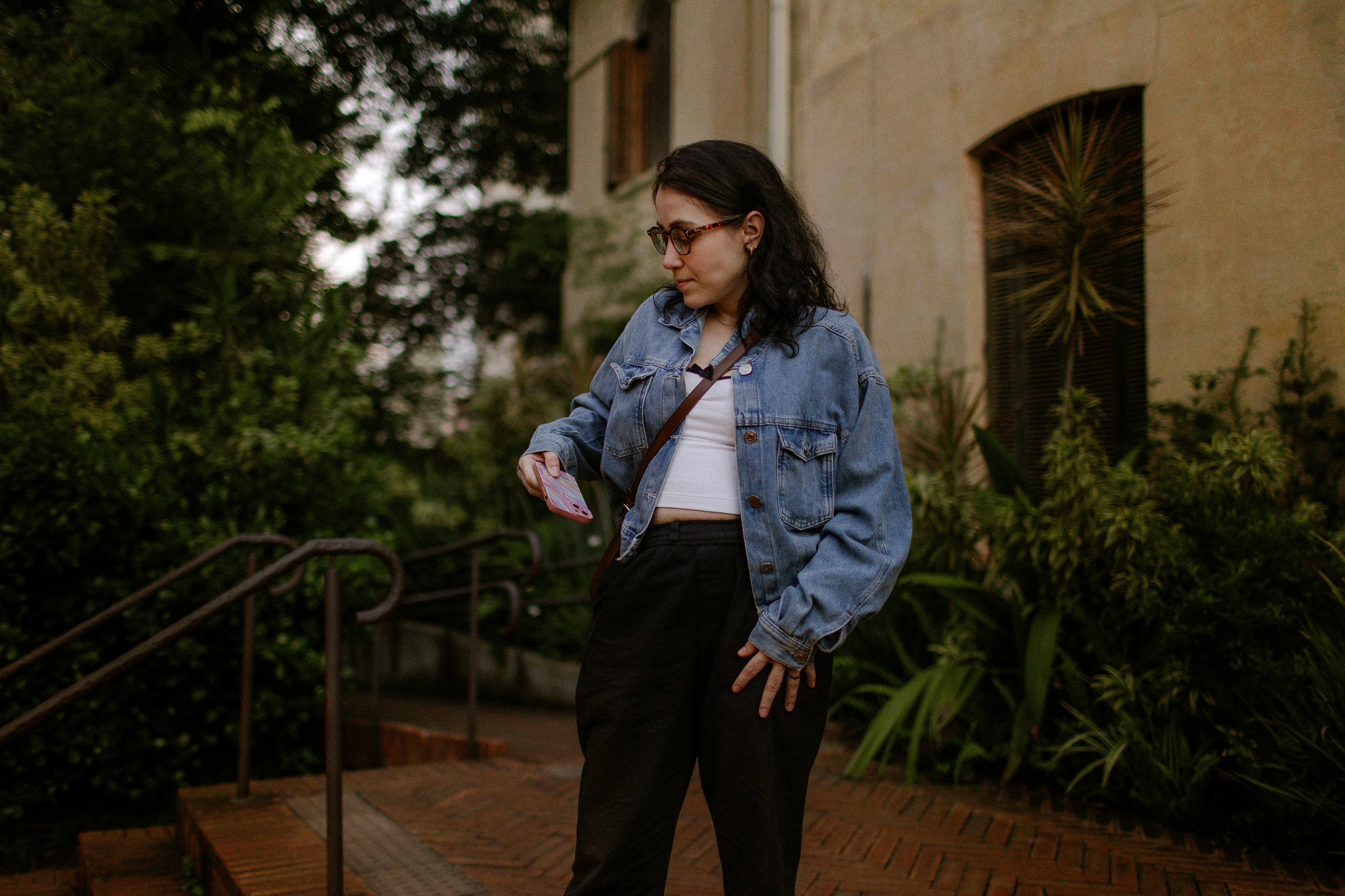 A woman in a denim jacket and glasses holding a phone outdoors