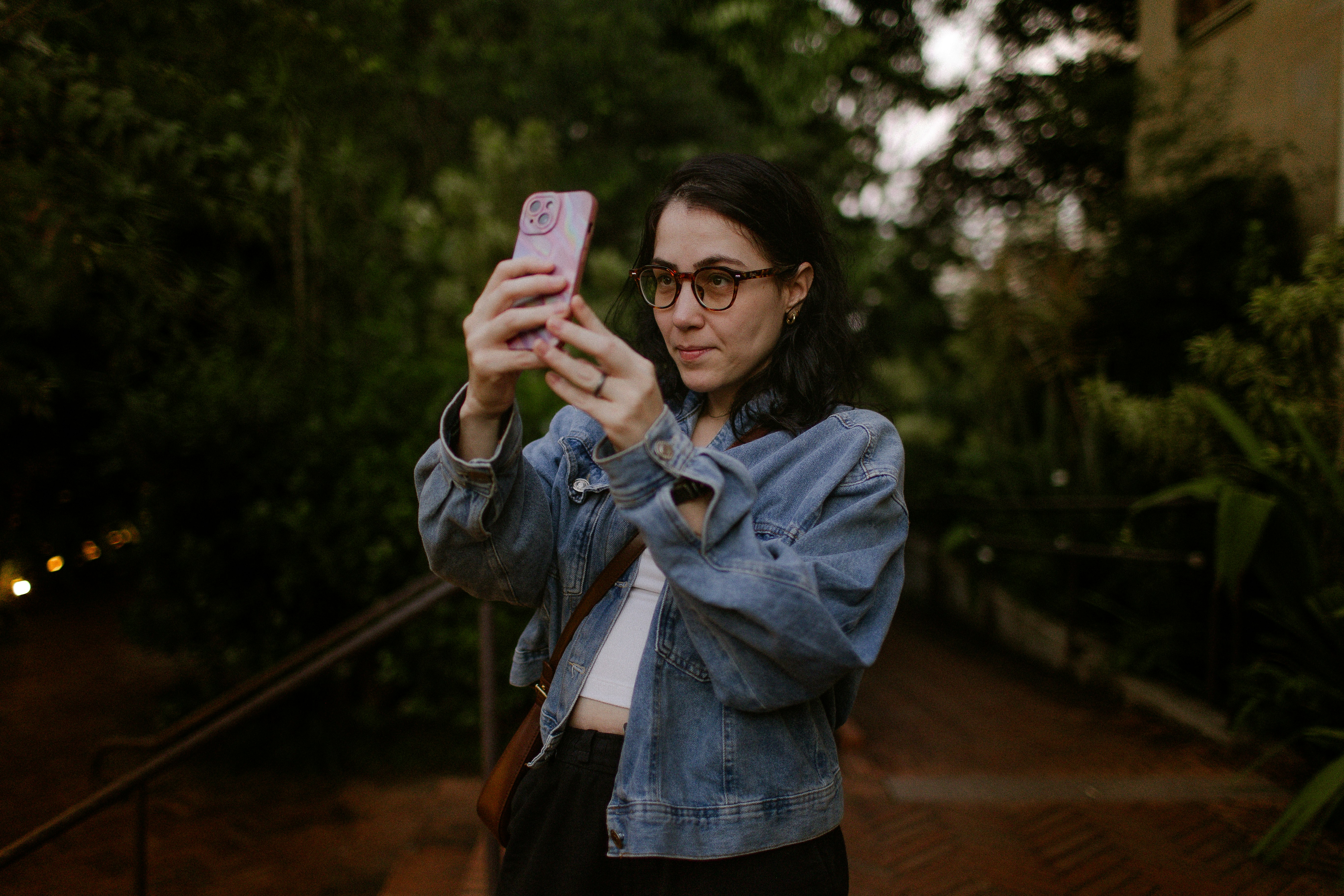 A woman with glasses holding a smartphone to take a photo outdoors