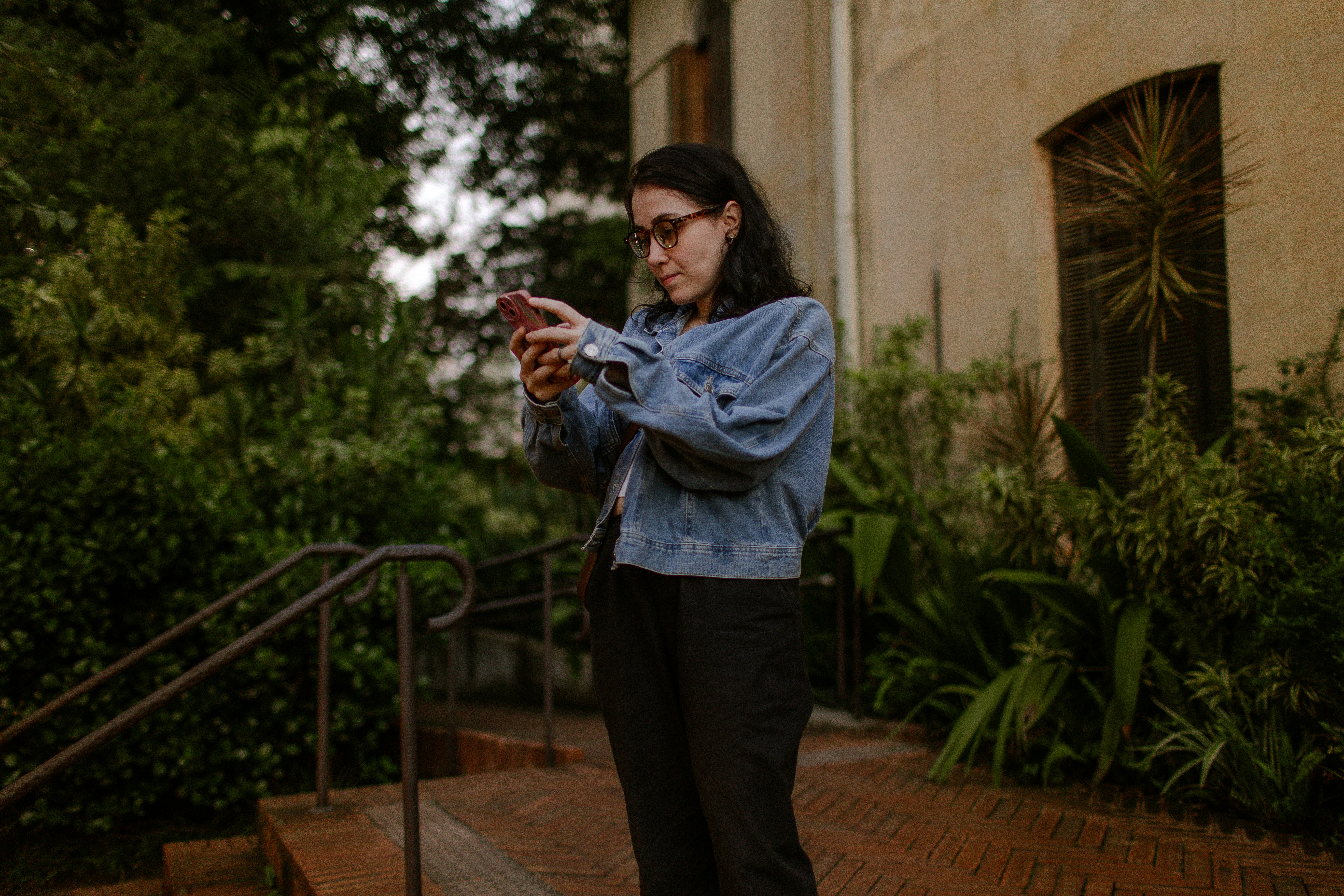 A woman in a denim jacket using a smartphone outdoors near lush greenery