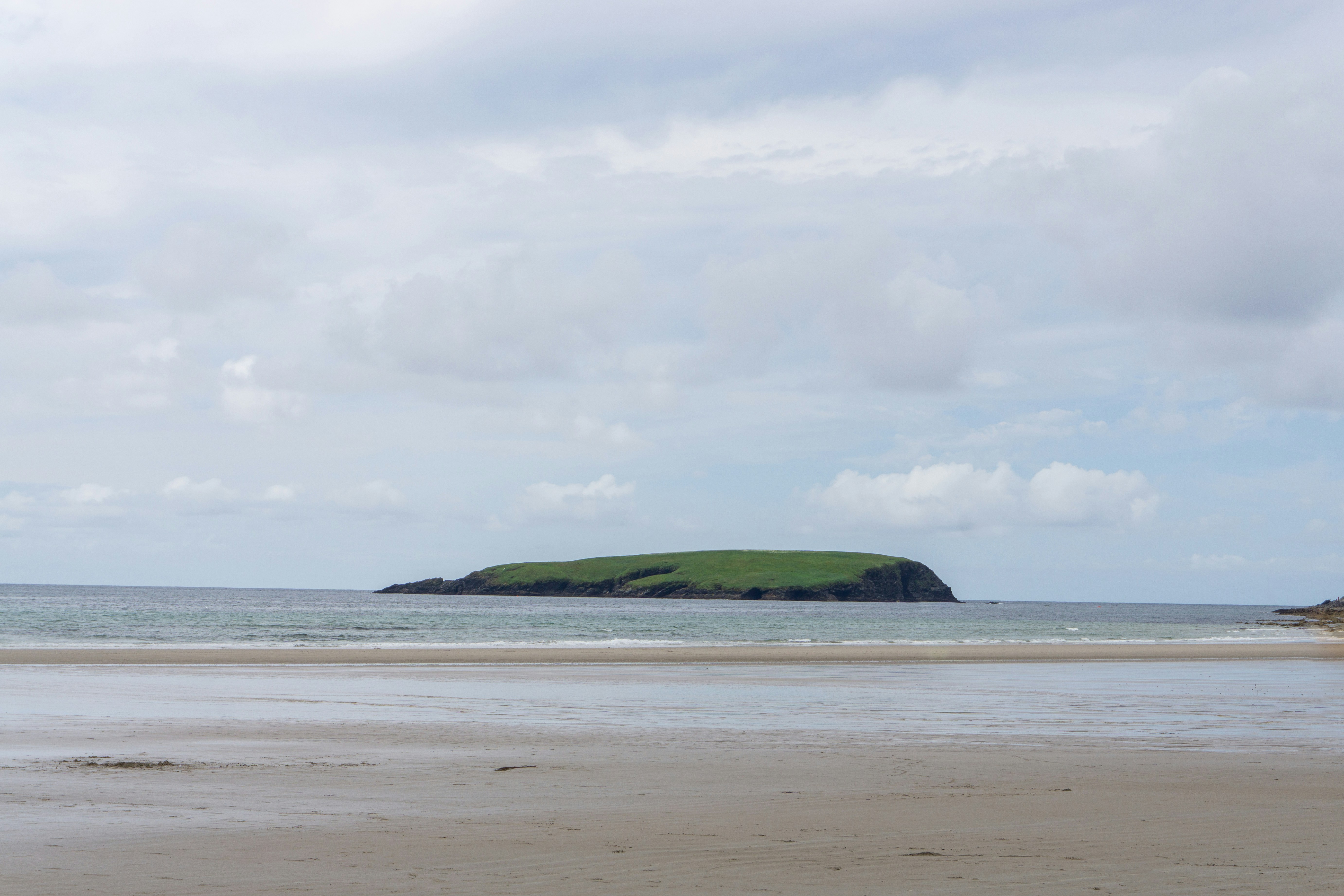 A small green island in the sea off a sandy beach under clouds