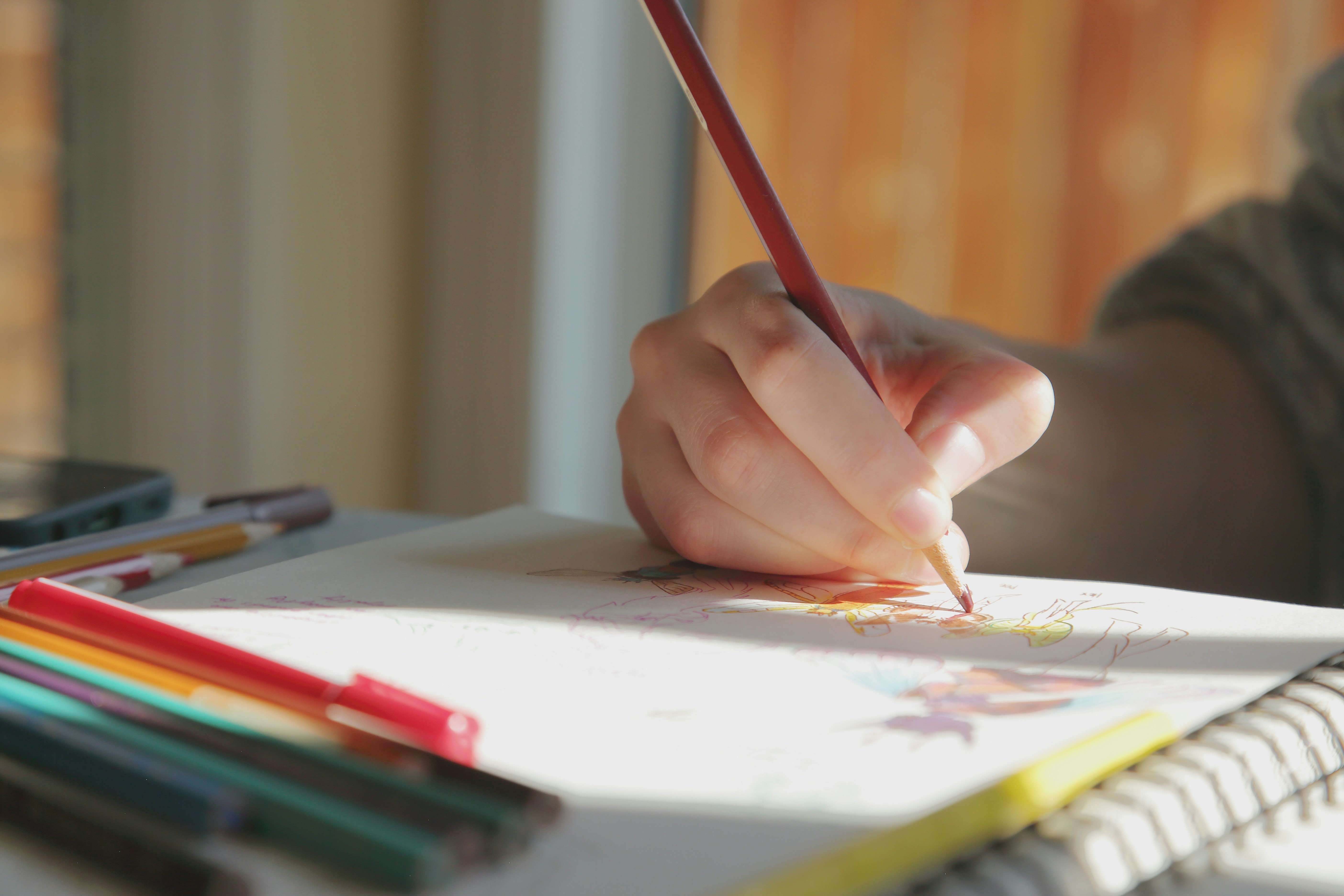 A hand holding a red pencil drawing on paper with colored pencils nearby