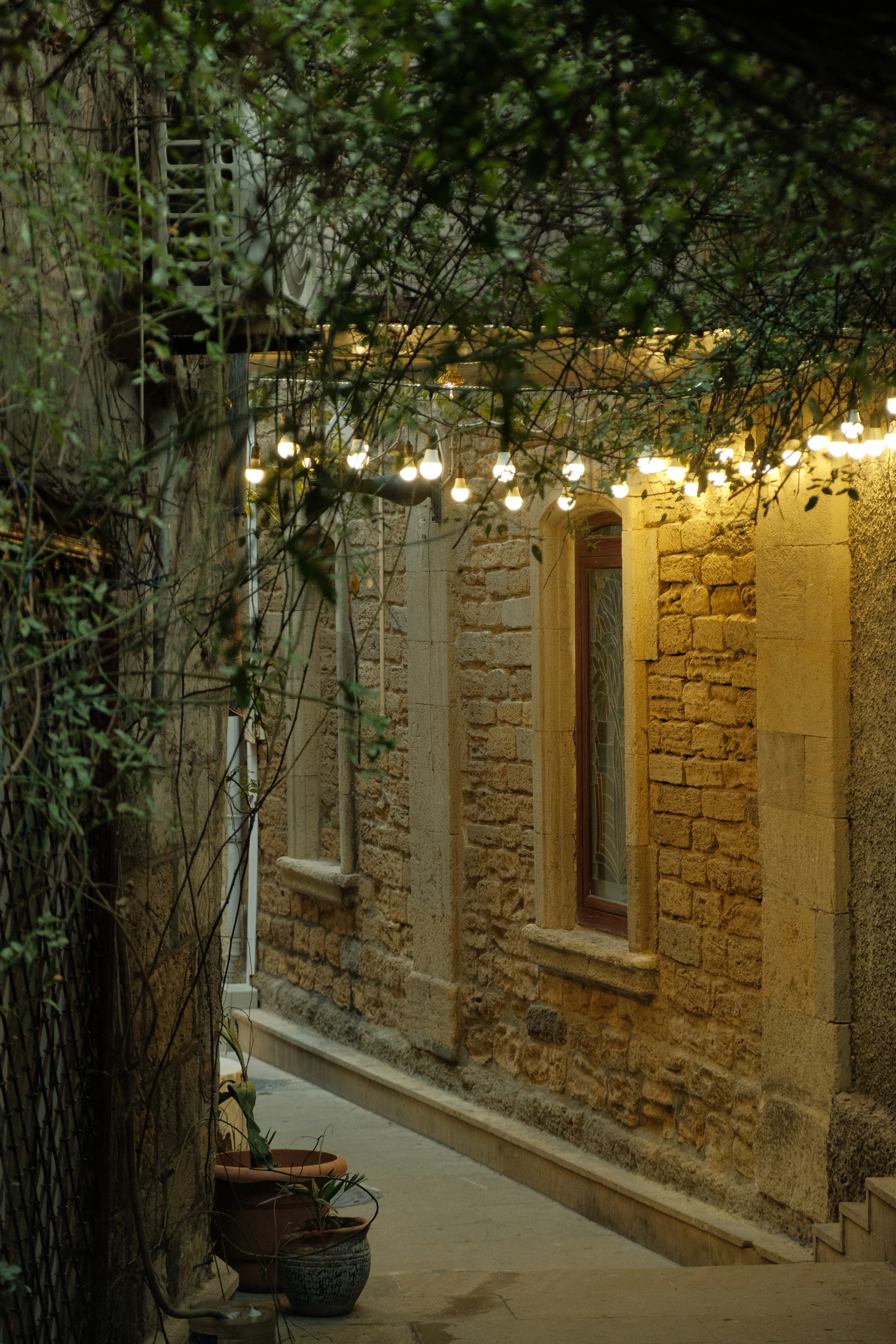 A narrow stone alley in Baku's Old City in the evening, strung overhead with warm filament bulbs that light the honey-coloured masonry, a tall arched window and terracotta pots on the step