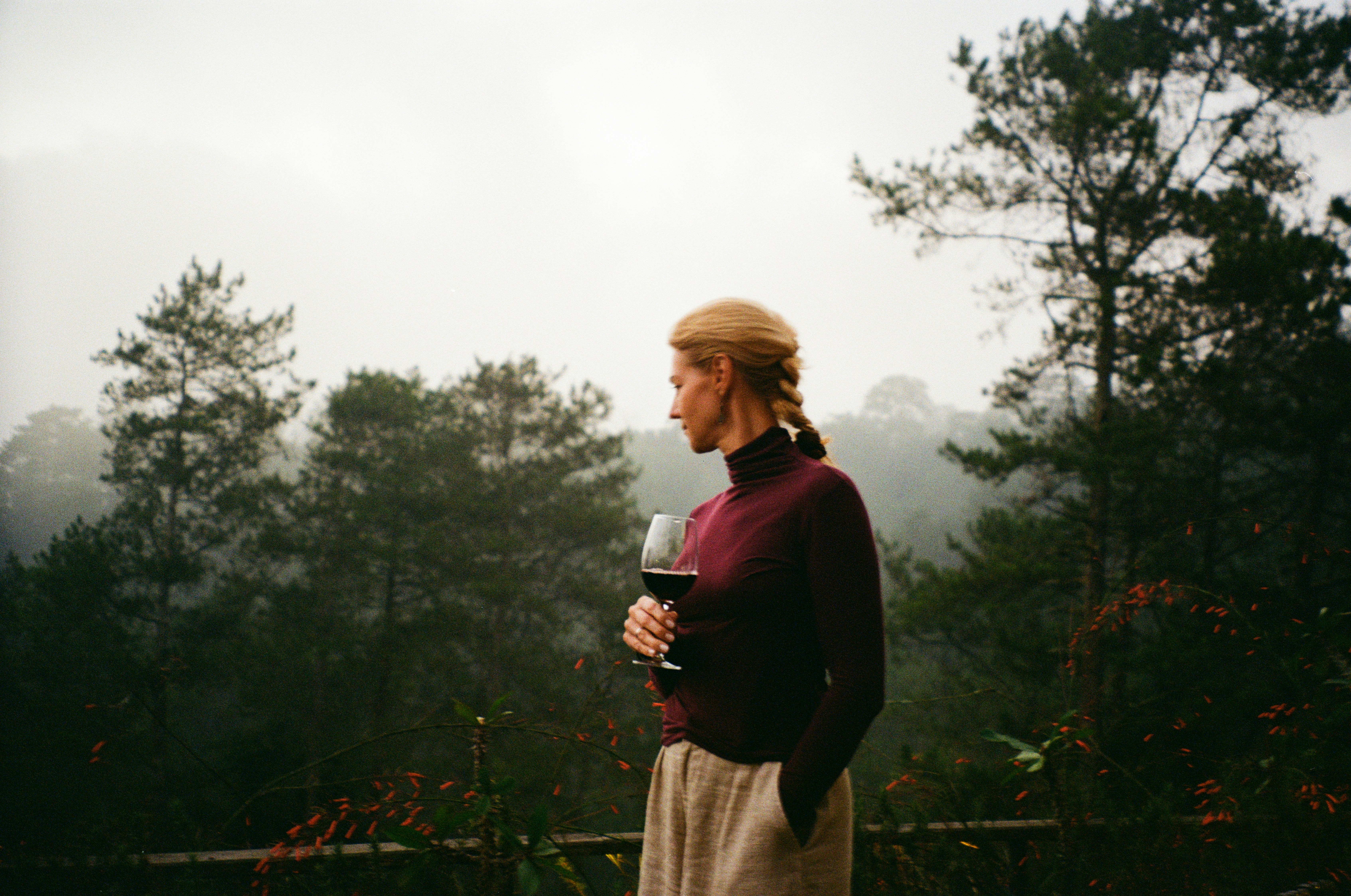 A woman in a burgundy turtleneck holding a wine glass in a misty forest