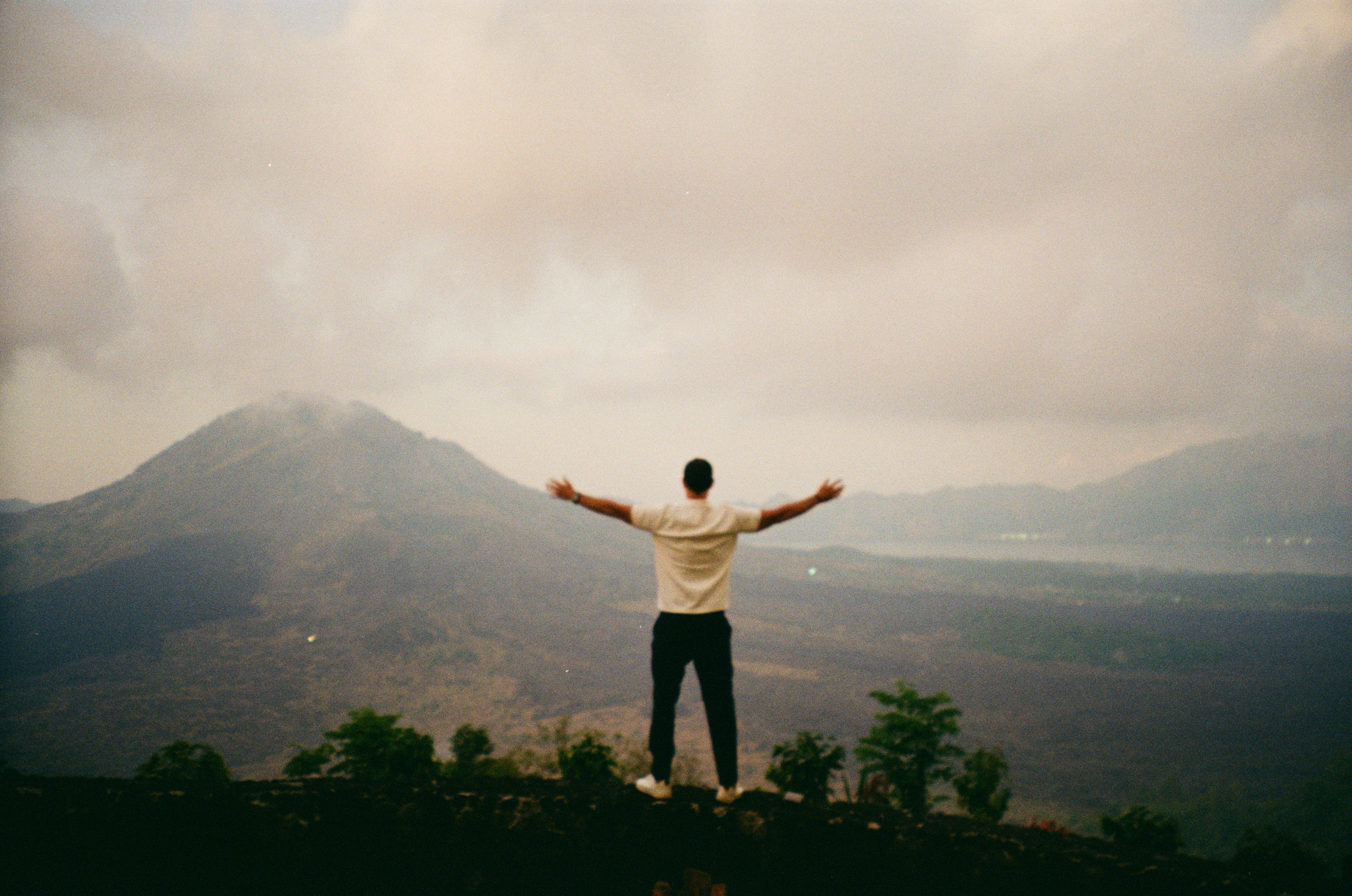 A person with arms outstretched overlooking a volcanic mountain landscape under a cloudy sky