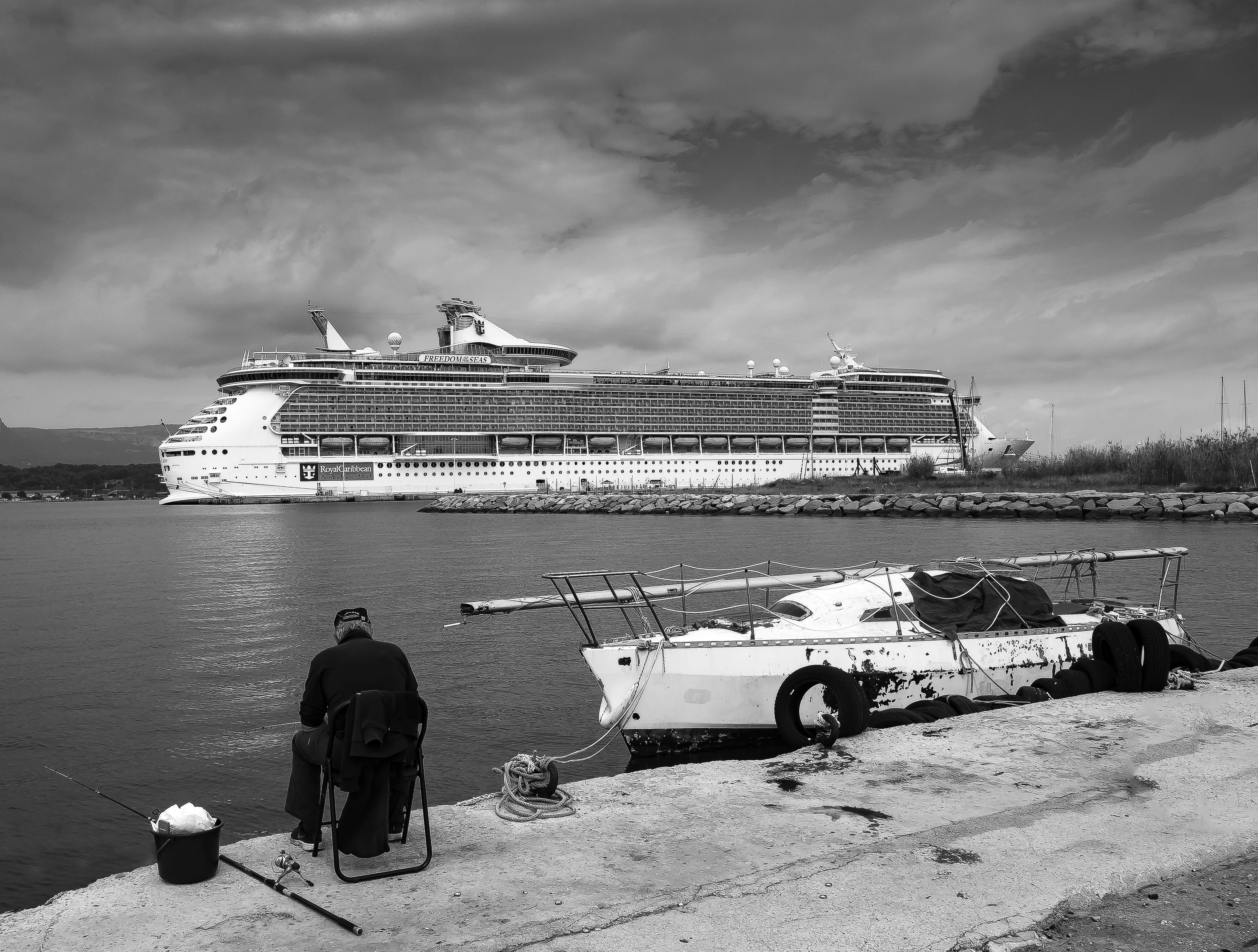 A person fishing from a concrete pier near a docked sailboat and cruise ship