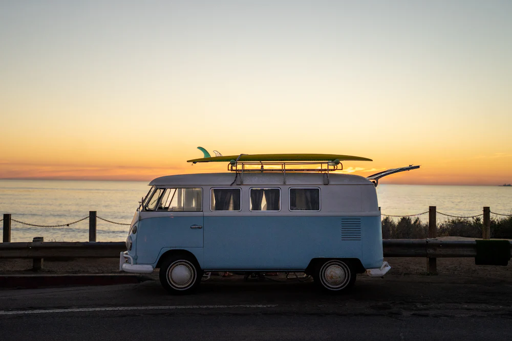 A pale blue vintage van parked by the ocean with a board on the roof.