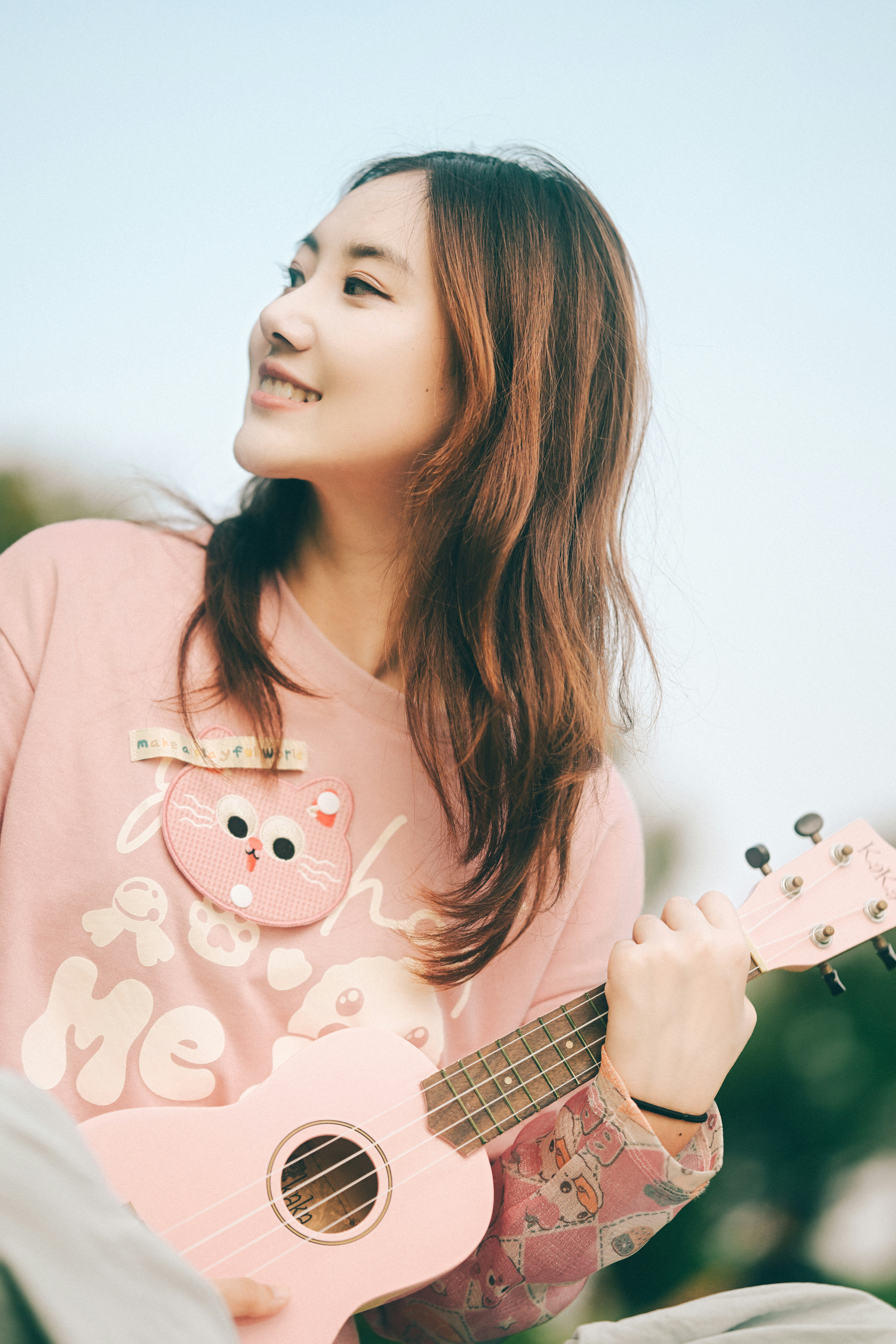 A woman in a pink shirt playing a pink ukulele outdoors