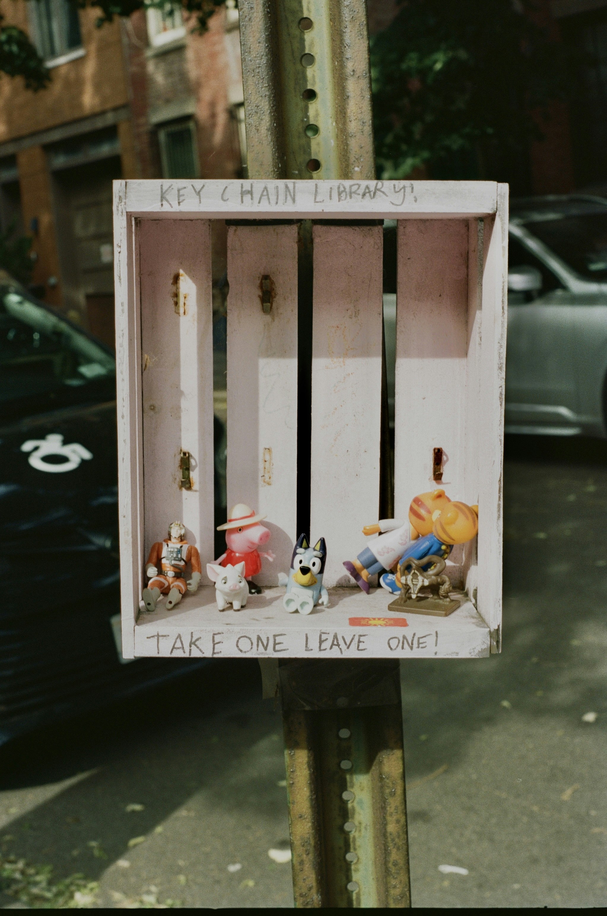 Small plastic figurines inside a wooden box labeled Key Chain Library on a post