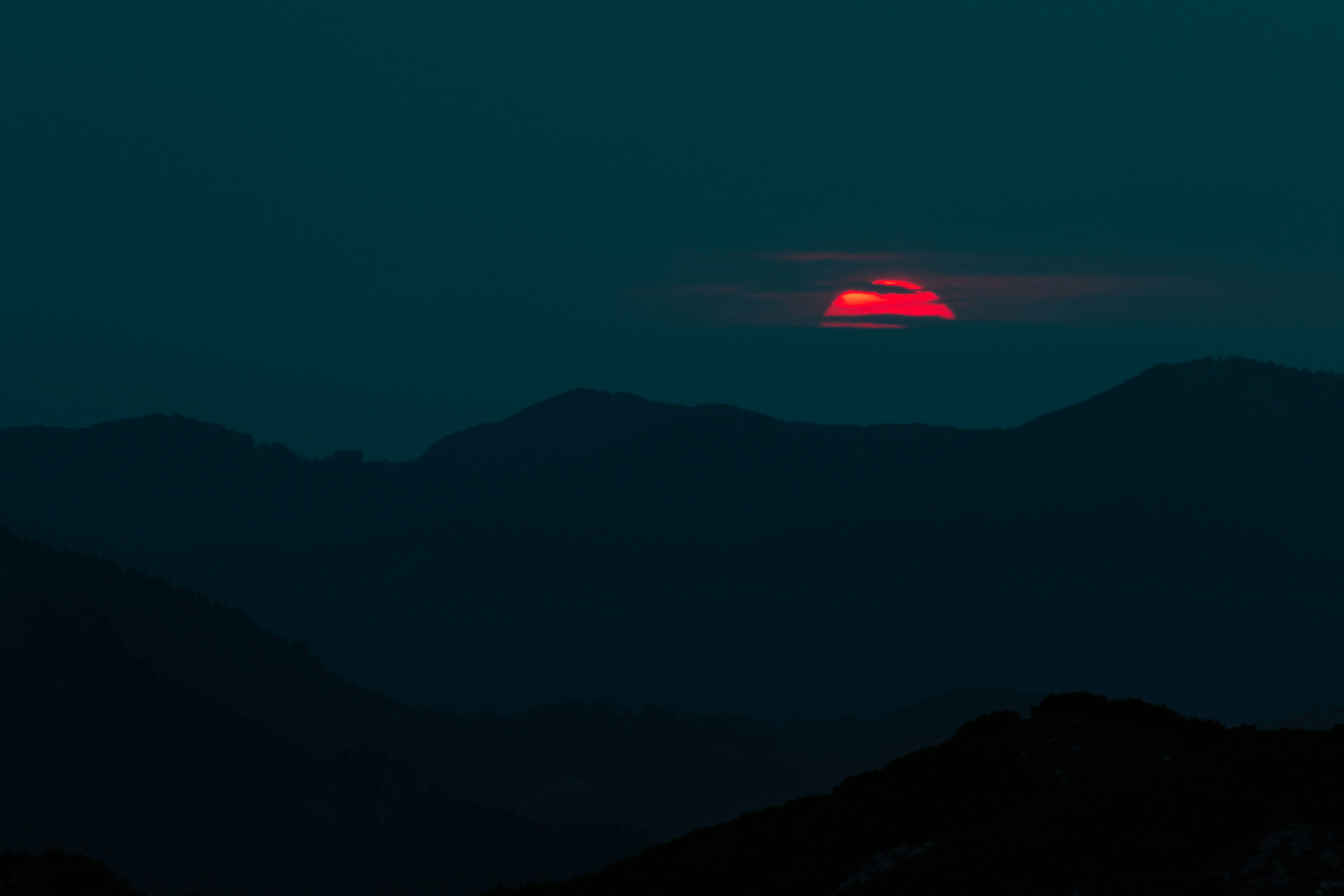 A bright red sun partially obscured by dark clouds above silhouetted mountain ridges