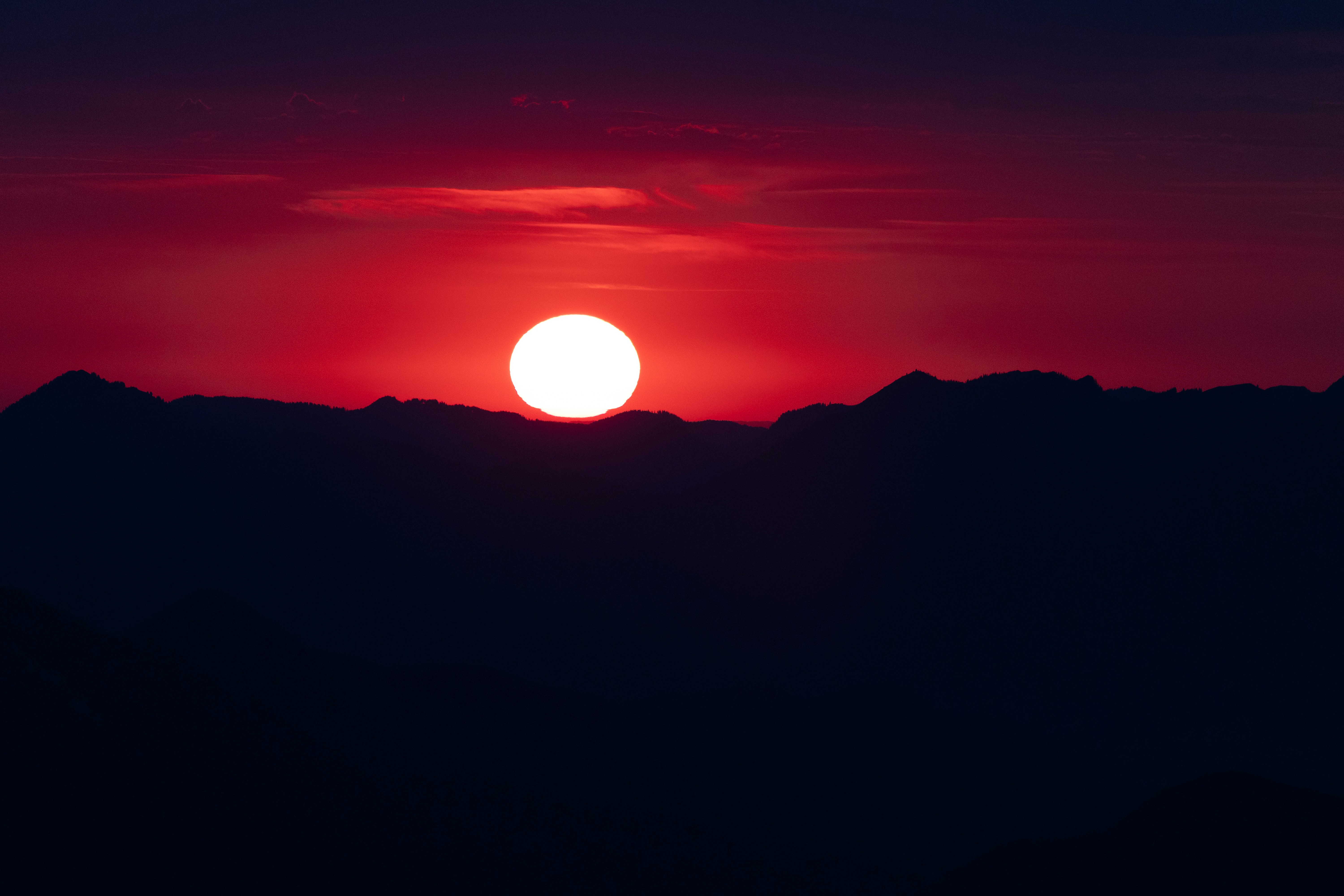 A bright white low sun above dark mountain silhouettes against a red sky