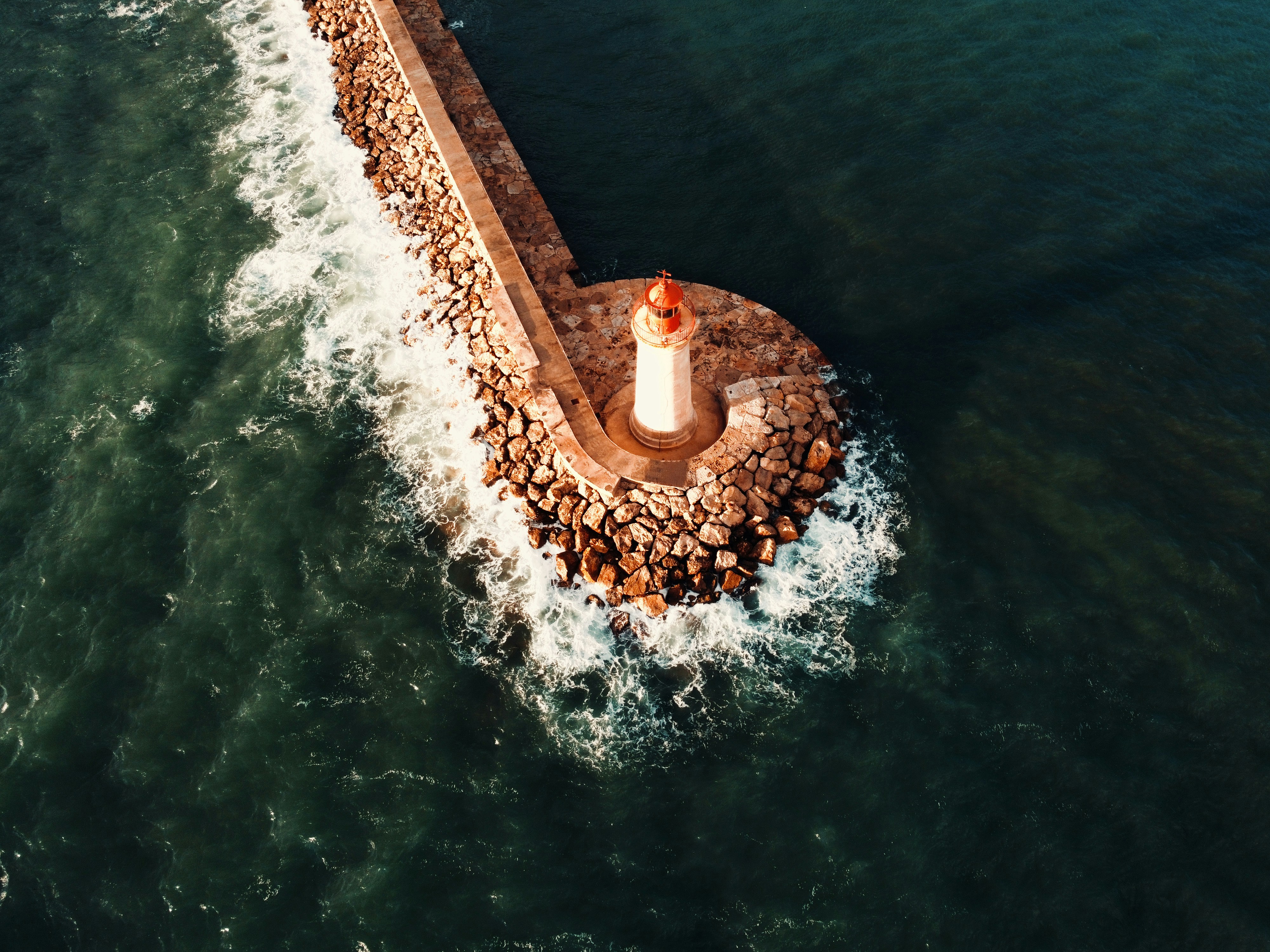 An aerial view of a lighthouse on a stone breakwater surrounded by dark water