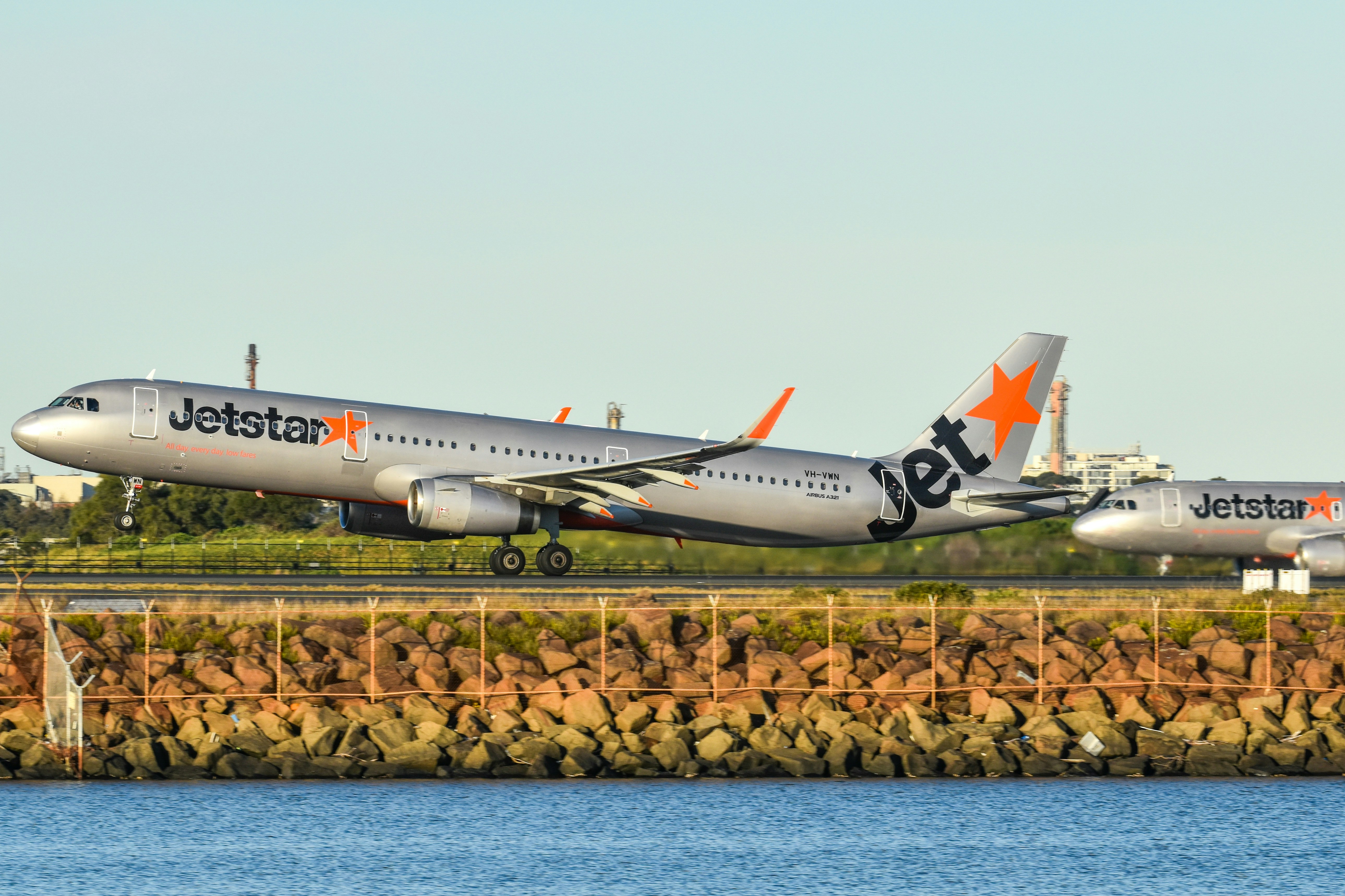 Two silver Jetstar passenger airplanes on a runway near a rocky shoreline