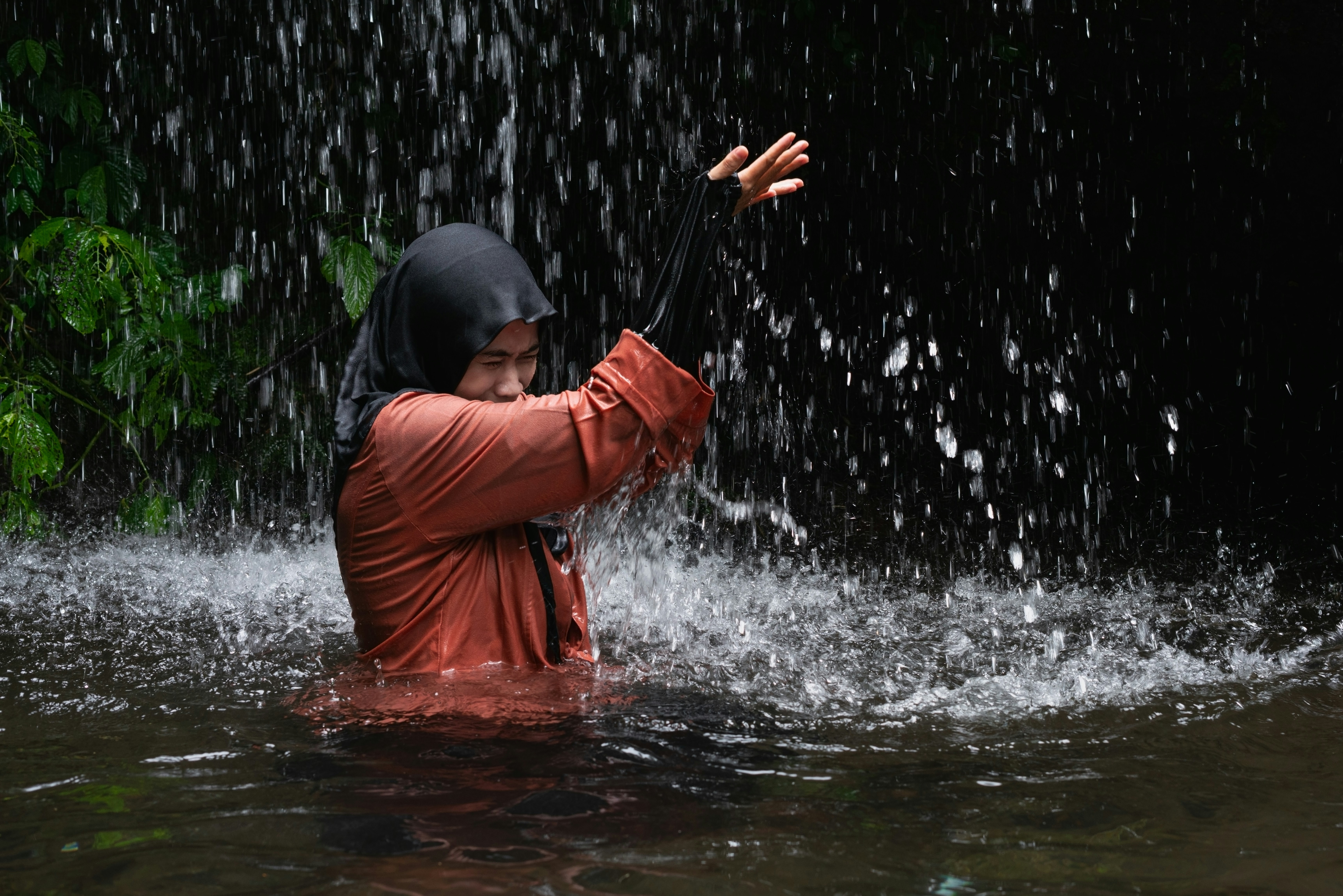 Una persona con hiyab y camiseta naranja de pie bajo una cascada