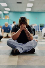 A person with tattoos sitting cross-legged on a floor with hands over their face
