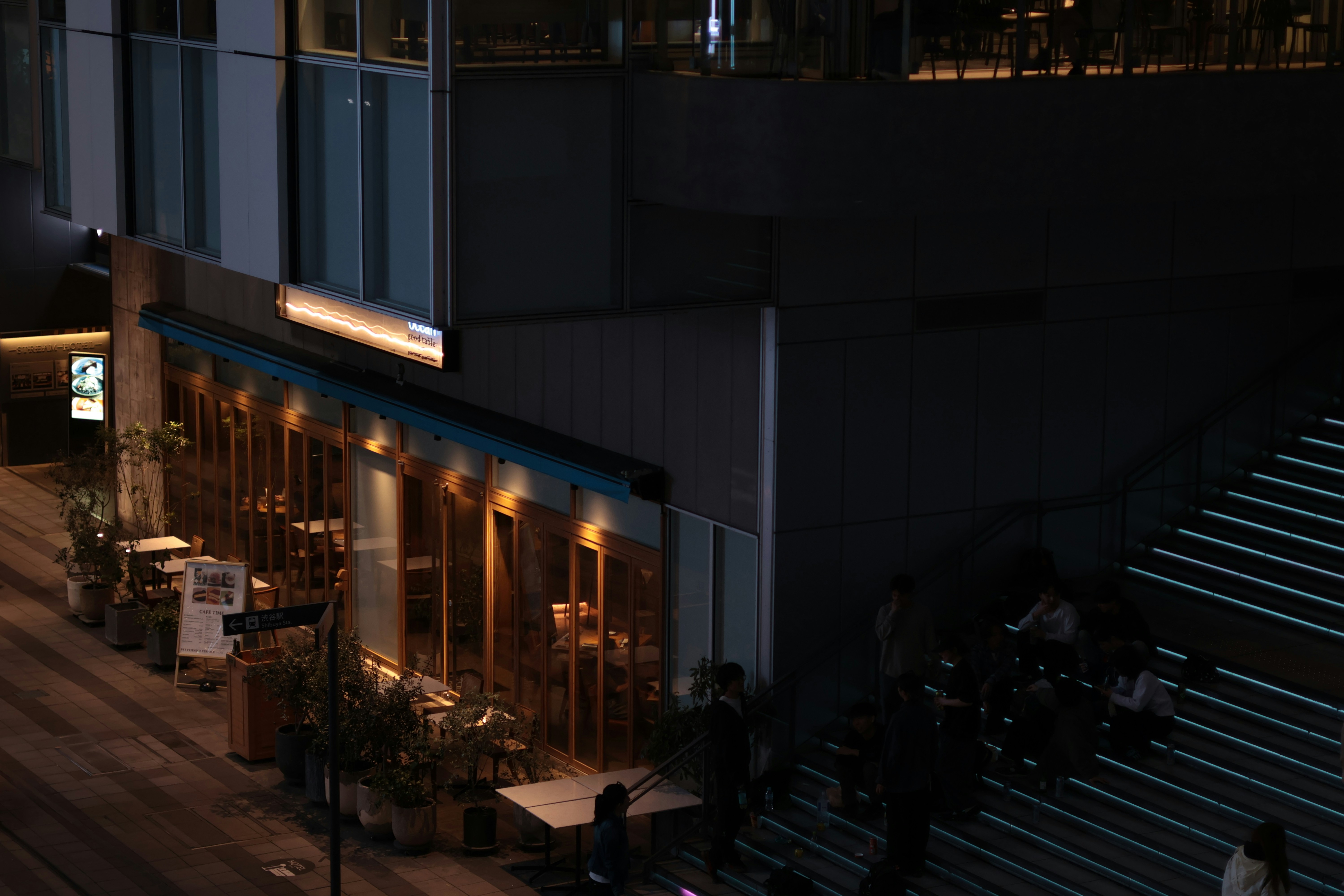 People sitting on illuminated outdoor stairs beside a restaurant with warm interior lighting