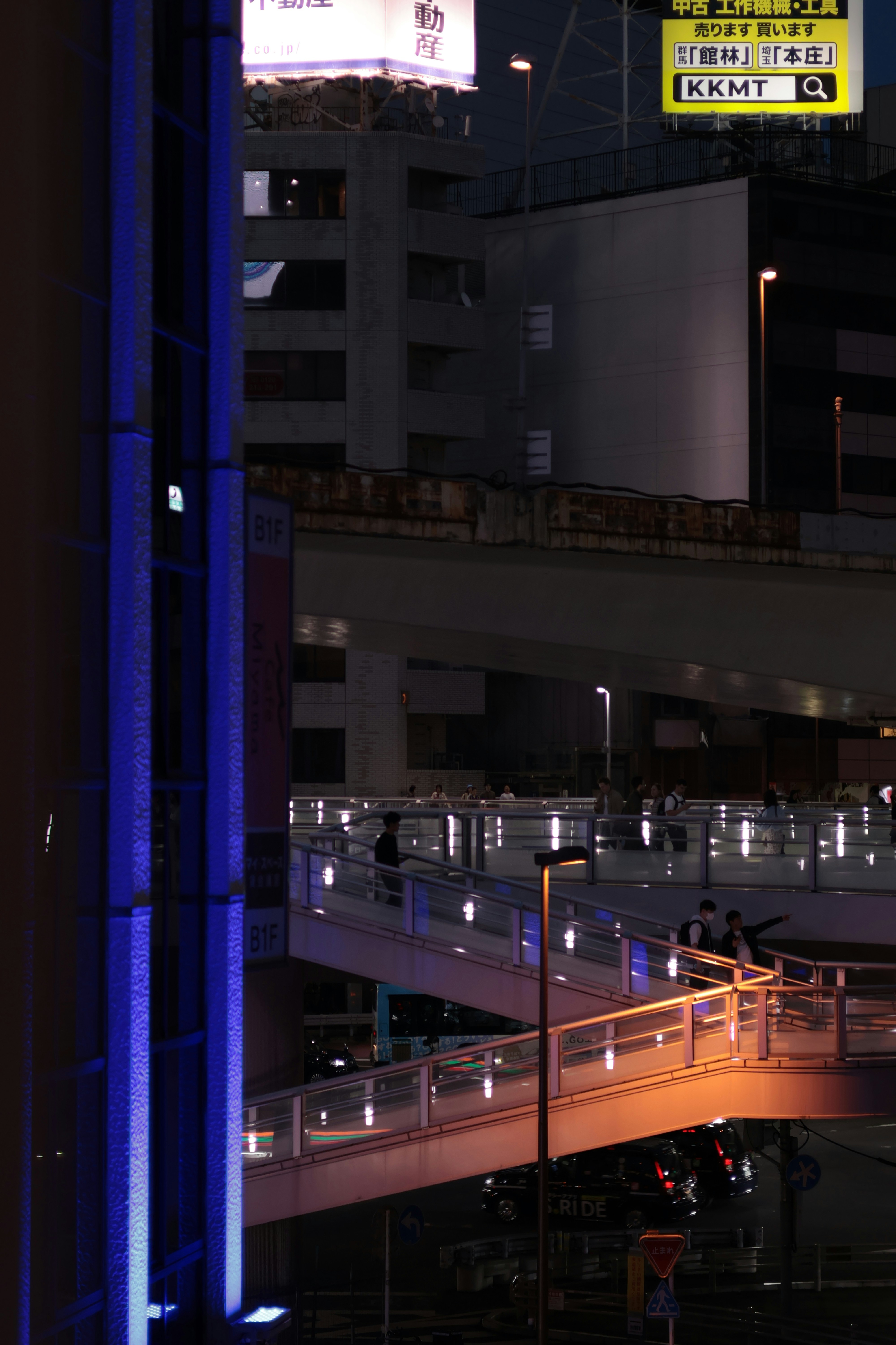 People walking on a multi-level pedestrian bridge at night with glowing lights