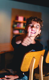 A person with curly hair giving a thumbs up in a wooden chair