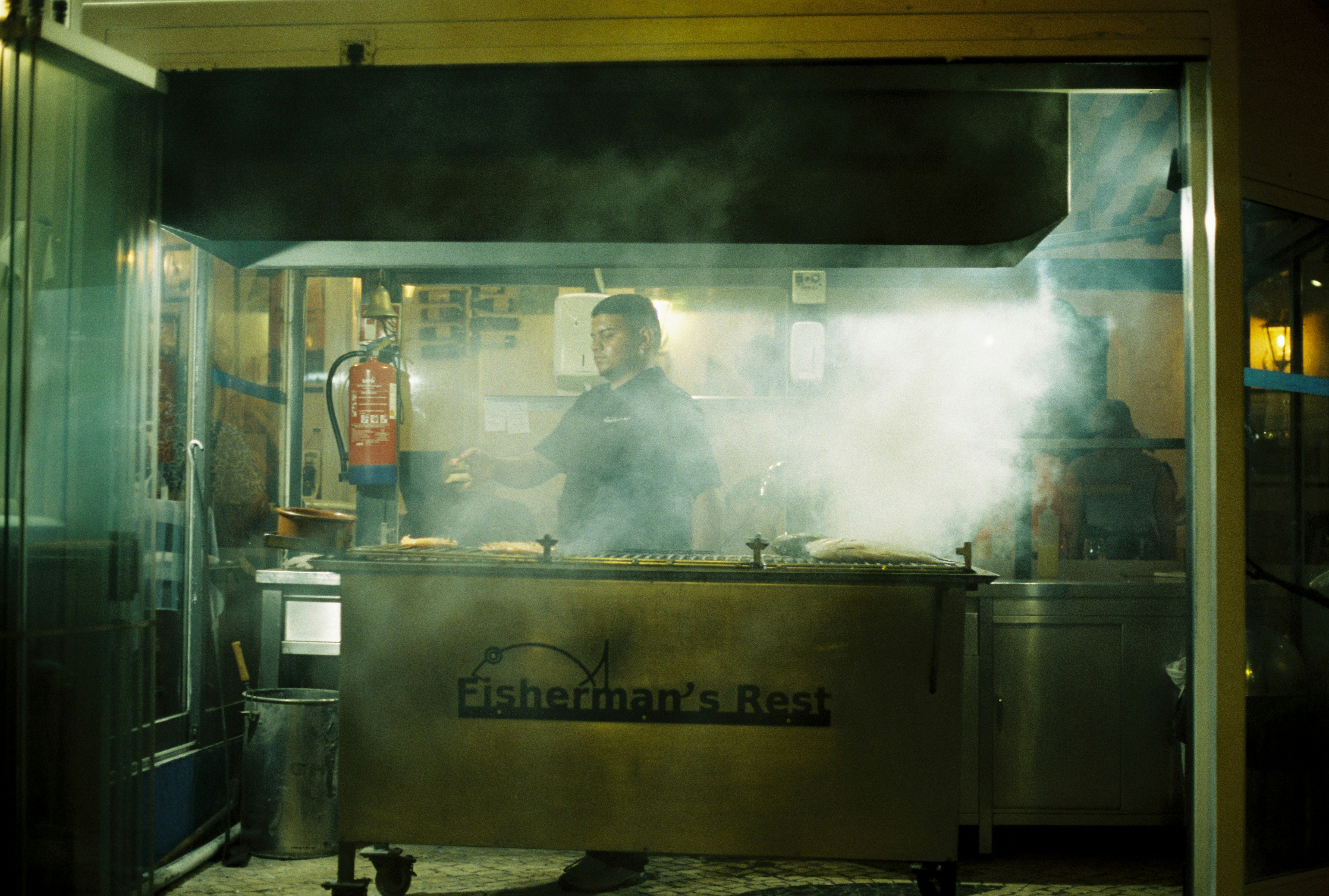 A cook tends a smoking charcoal grill outside a stall called Fisherman's Rest at night. - 相关推荐