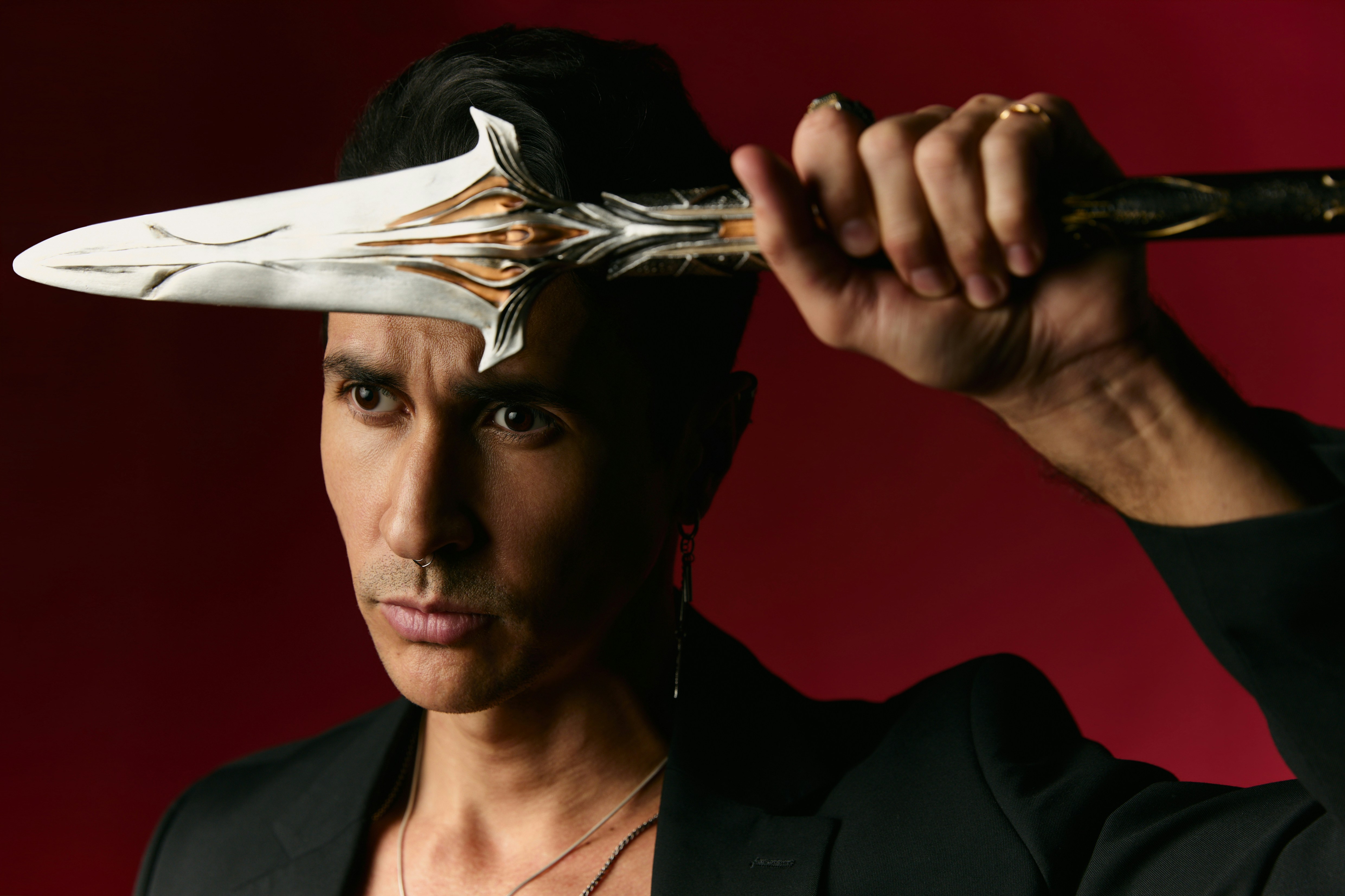 A person in a black blazer holding a decorative metal dagger near their forehead