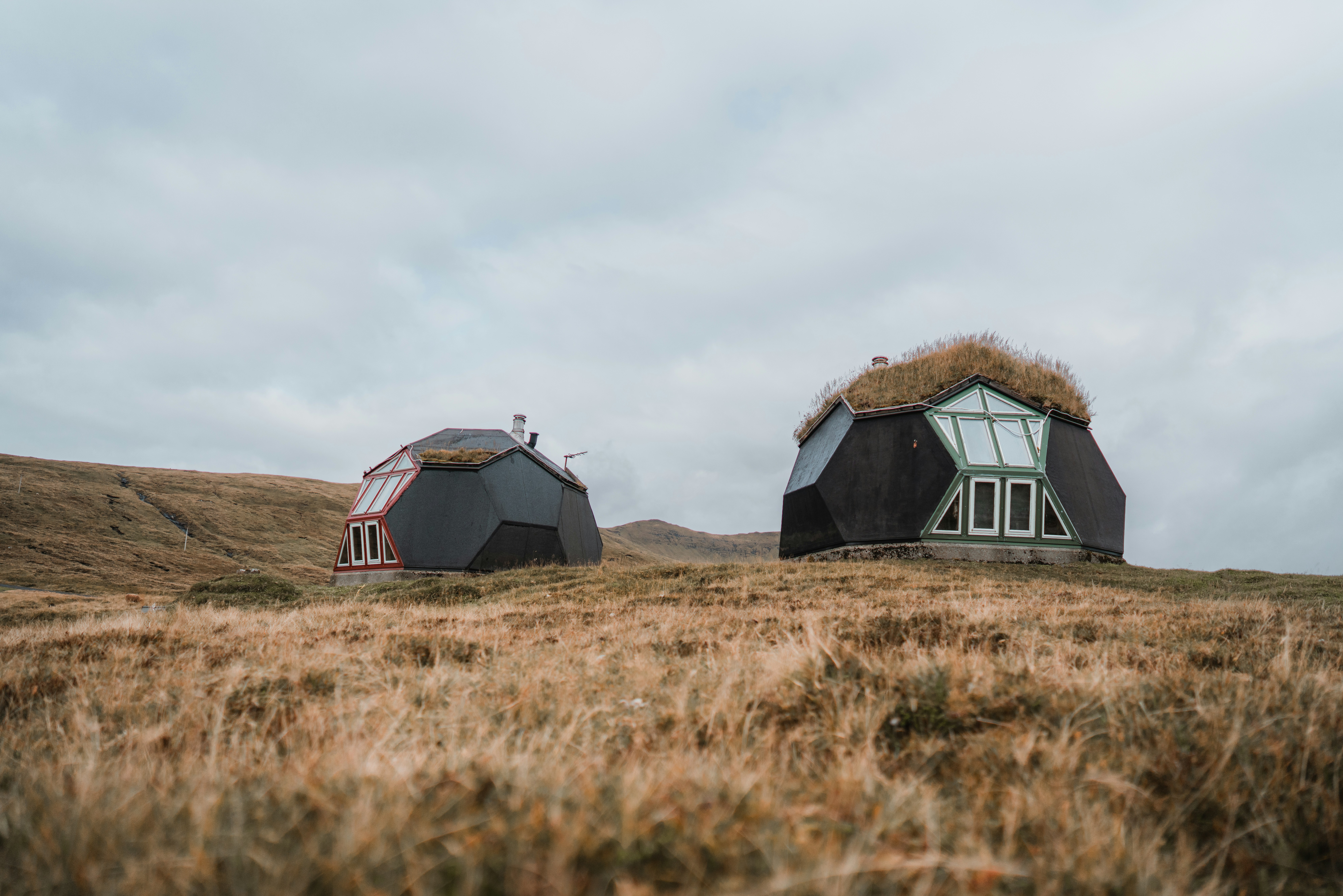 Two geometric cabins with grass roofs on a dry grassy hillside under cloudy skies
