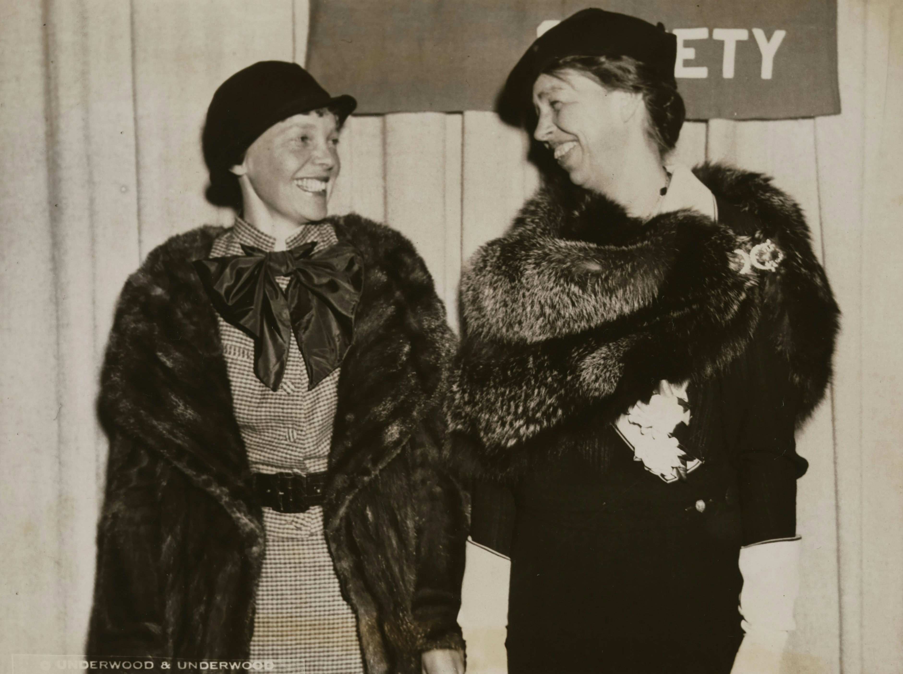 Two women in hats and fur coats smiling at each other indoors