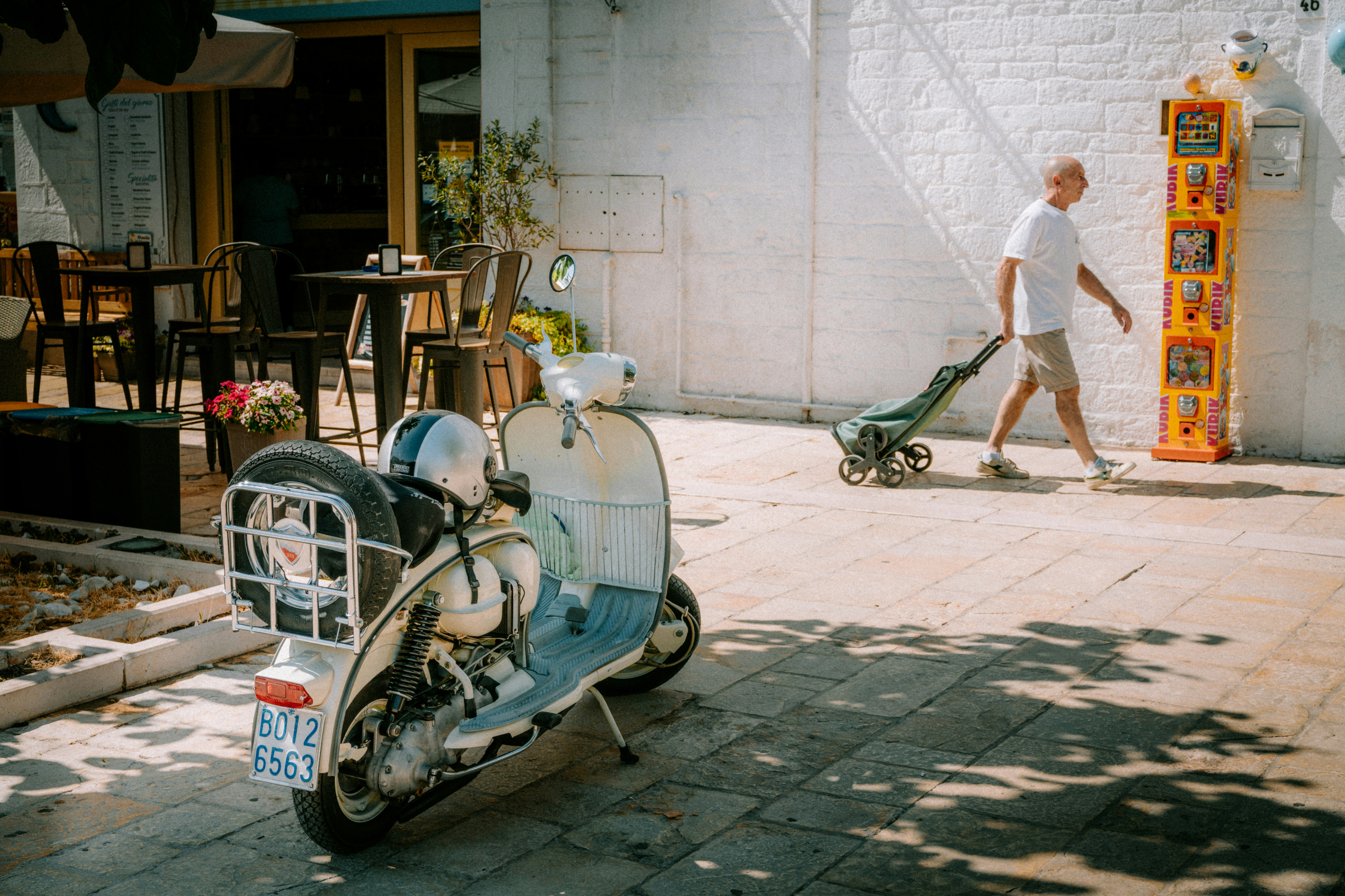 A man walking past a parked white scooter on a sunlit stone street