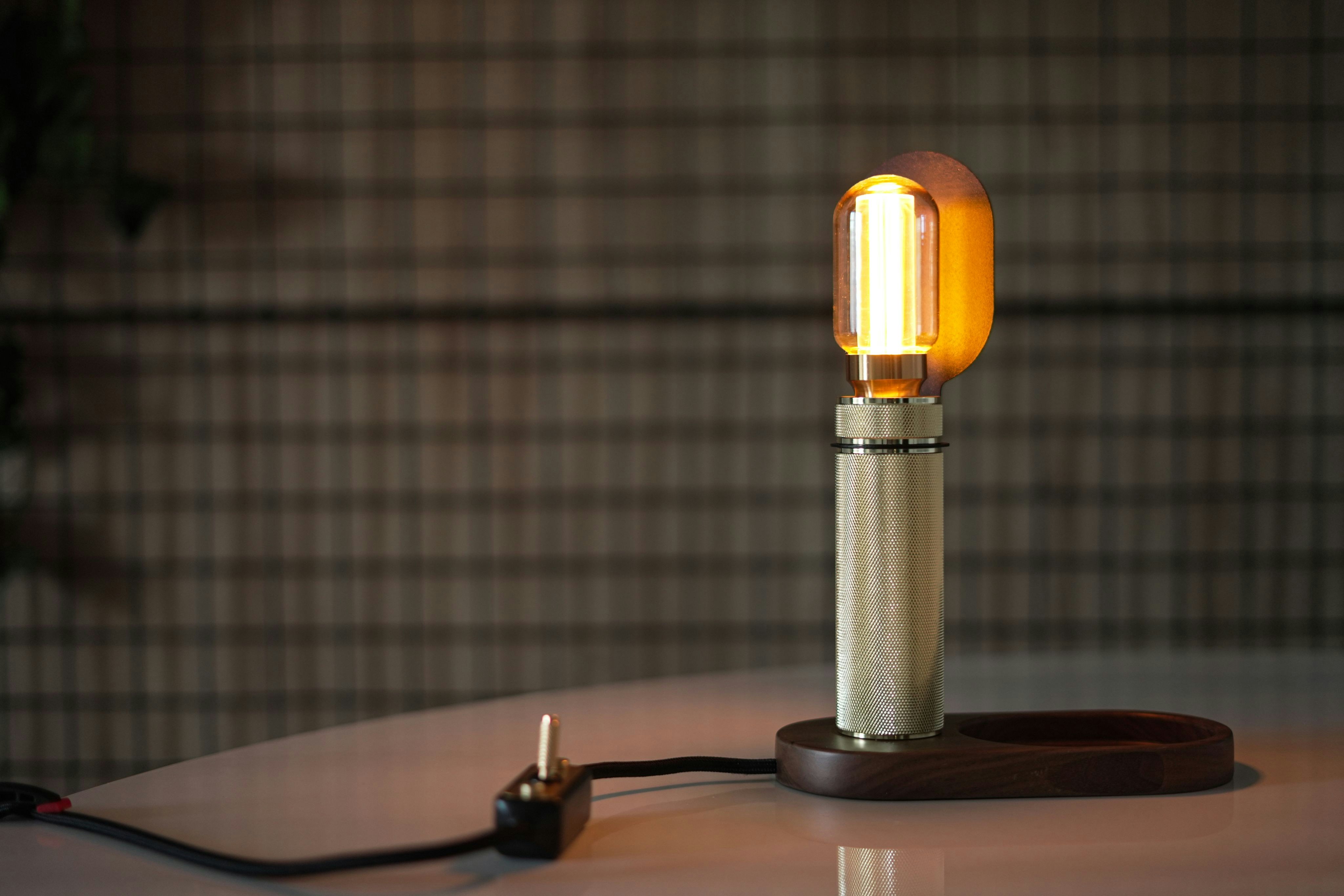 A lit cylindrical brass lamp with a wooden base on a white surface